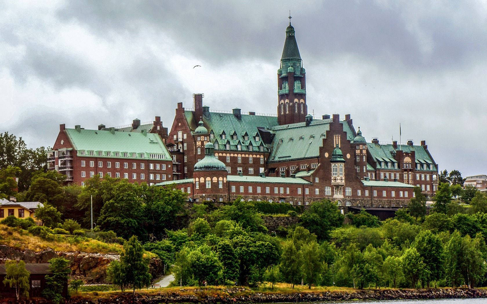 Sweden 桌布 壁紙 — 一座宏偉的歷史磚砌建築，擁有綠色銅屋頂，坐落在鬱蔥的山坡上，俯瞰著多雲天空下的水域。