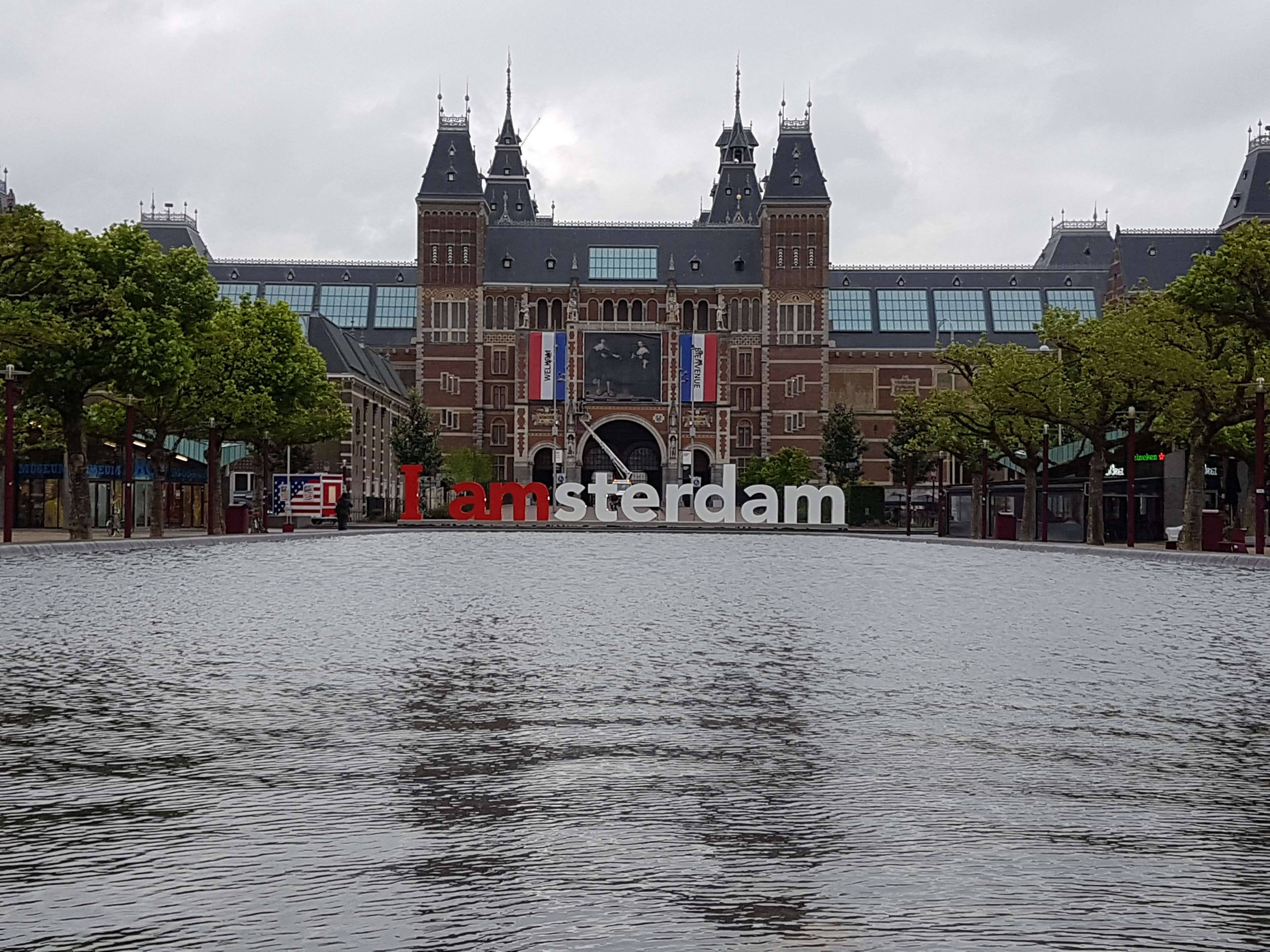 Amsterdam 桌布 壁紙 — 一座宏偉的歷史阿姆斯特丹建築矗立在反射水景後面，前方有一個大型「I amsterdam」標誌。