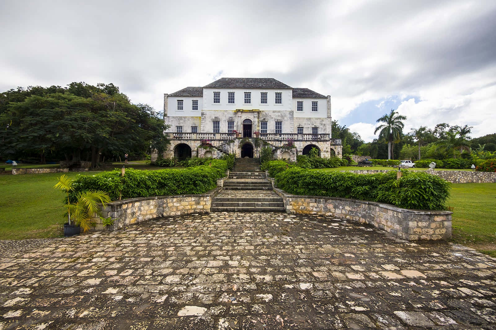Jamaica 桌布 壁紙 — 一座宏偉的殖民地風格大宅，白色牆壁和石階，矗立在多雲的天空下，四周環繞著茂密的綠色草坪和熱帶樹木。