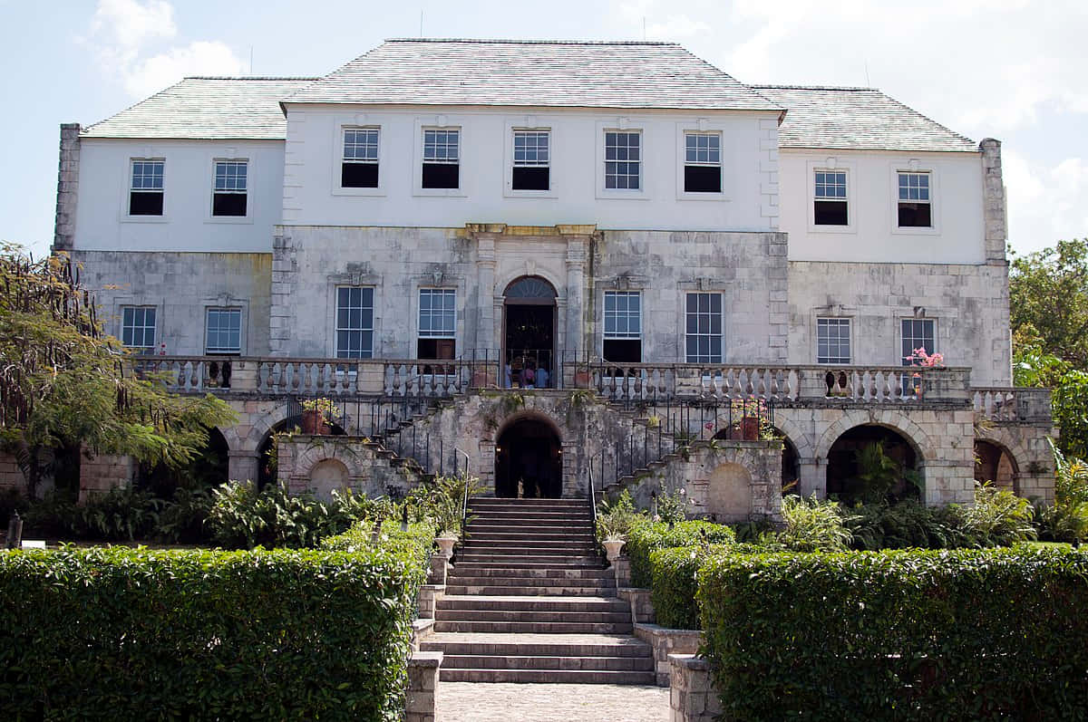 Jamaica 桌布 壁紙 — 一座宏偉的殖民風格大宅，擁有石階梯和拱形步道，周圍環繞著茂密的綠色樹籬，天空明亮。
