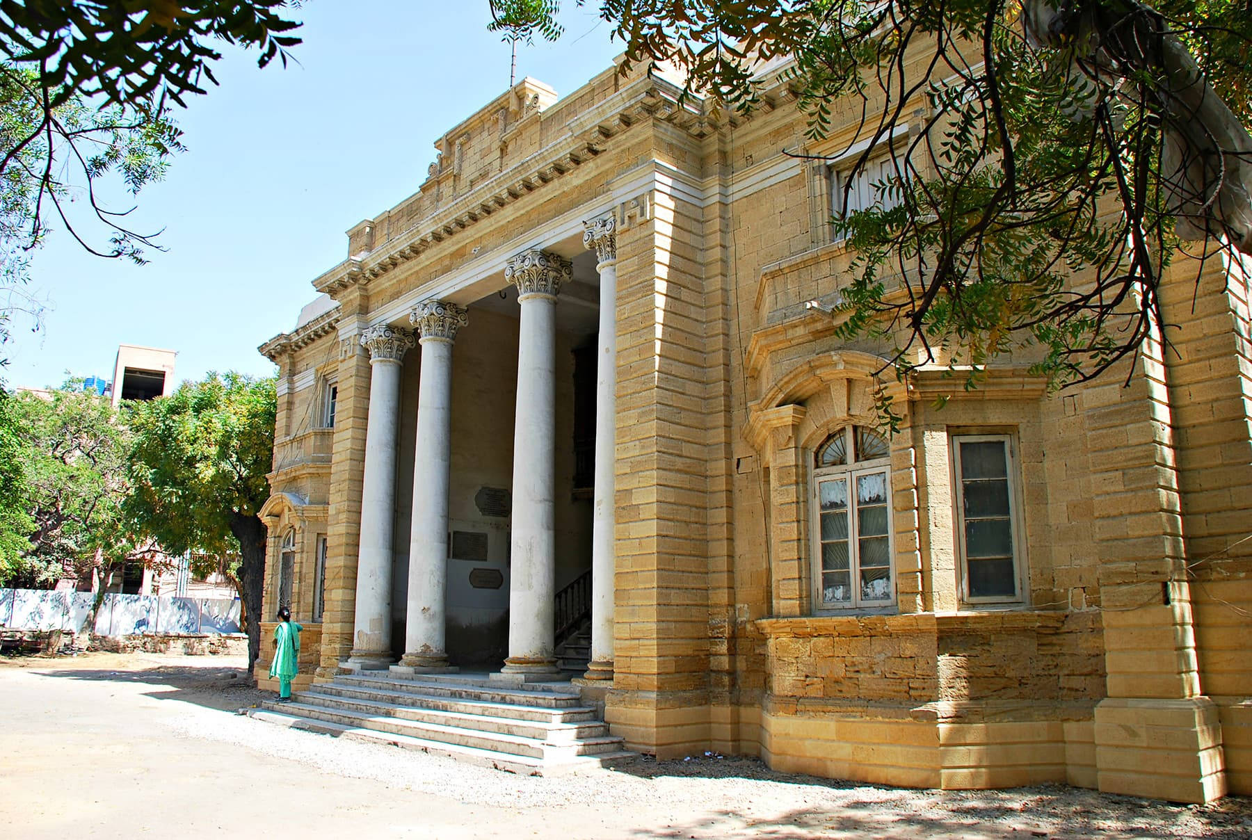 Karachi 桌布 壁紙 — 一座宏偉的殖民風格建築，擁有石柱和拱形窗戶，在明媚陽光下矗立，被枝葉茂密的樹枝環繞。