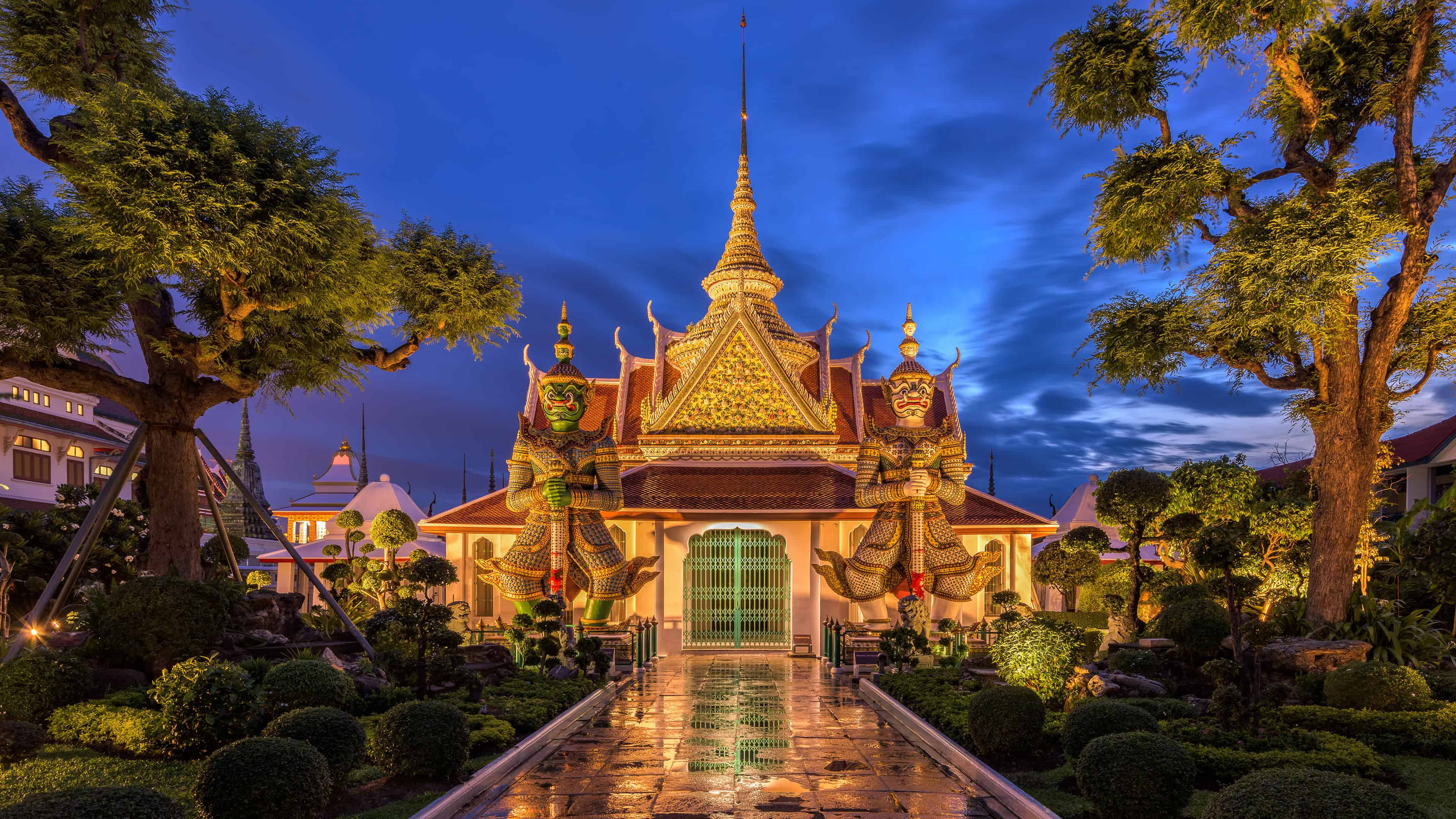 Bangkok 桌布 壁紙 — 一座宏偉的泰國寺廟在黃昏時分被照亮，金色尖頂和守護神雕像兩旁是茂密的樹木和修剪整齊的花園。