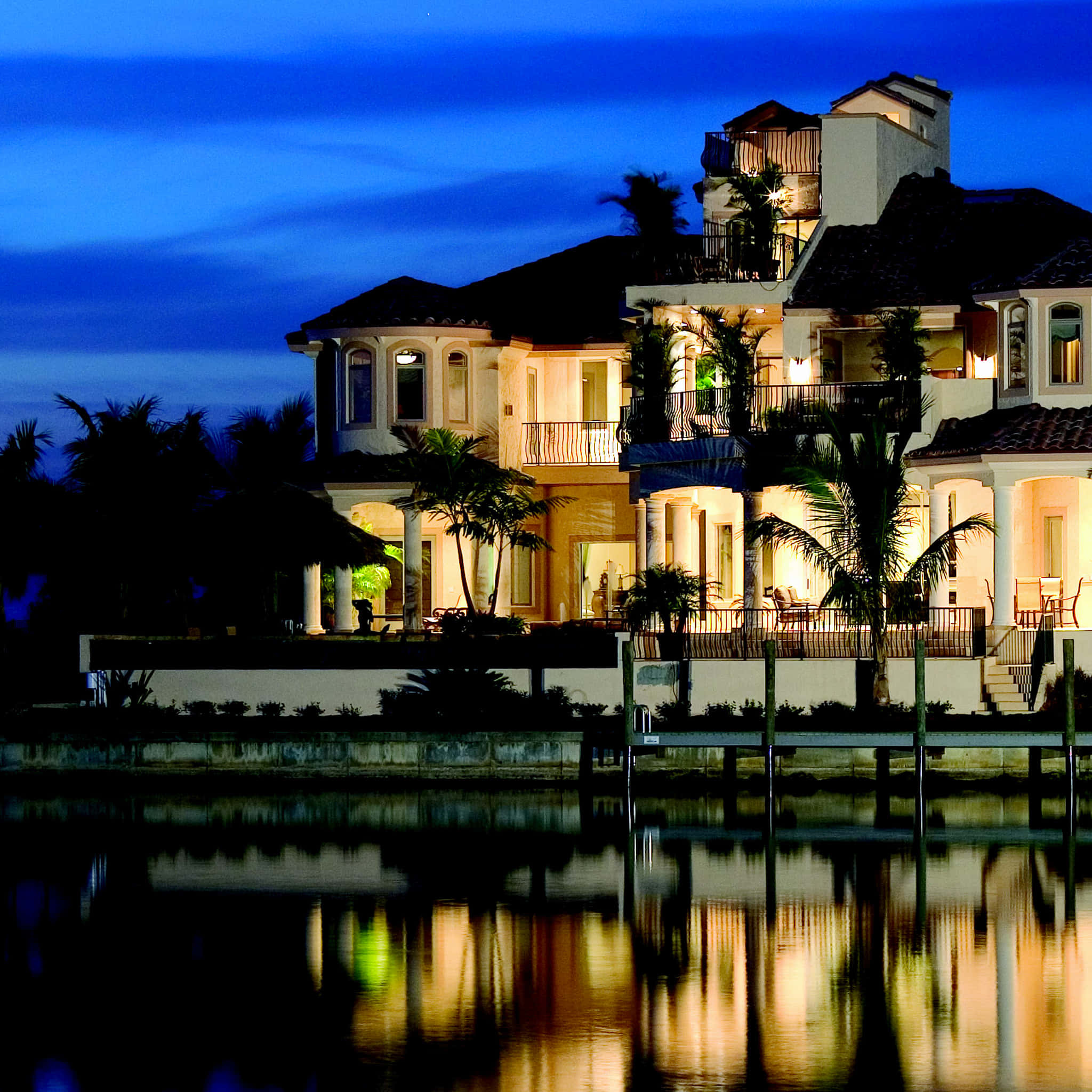 Mansion 桌布 壁紙 — 一座宏偉的海濱豪宅在黃昏時溫暖地發光，棕櫚樹及其燈光倒映在平靜的水面上。