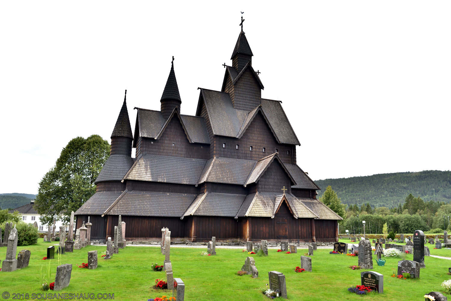 Norway 桌布 壁紙 — 一座宏偉的深色木造木板教堂，有多個尖頂，莊嚴地矗立在充滿活力的綠色墓地中，天空多雲。