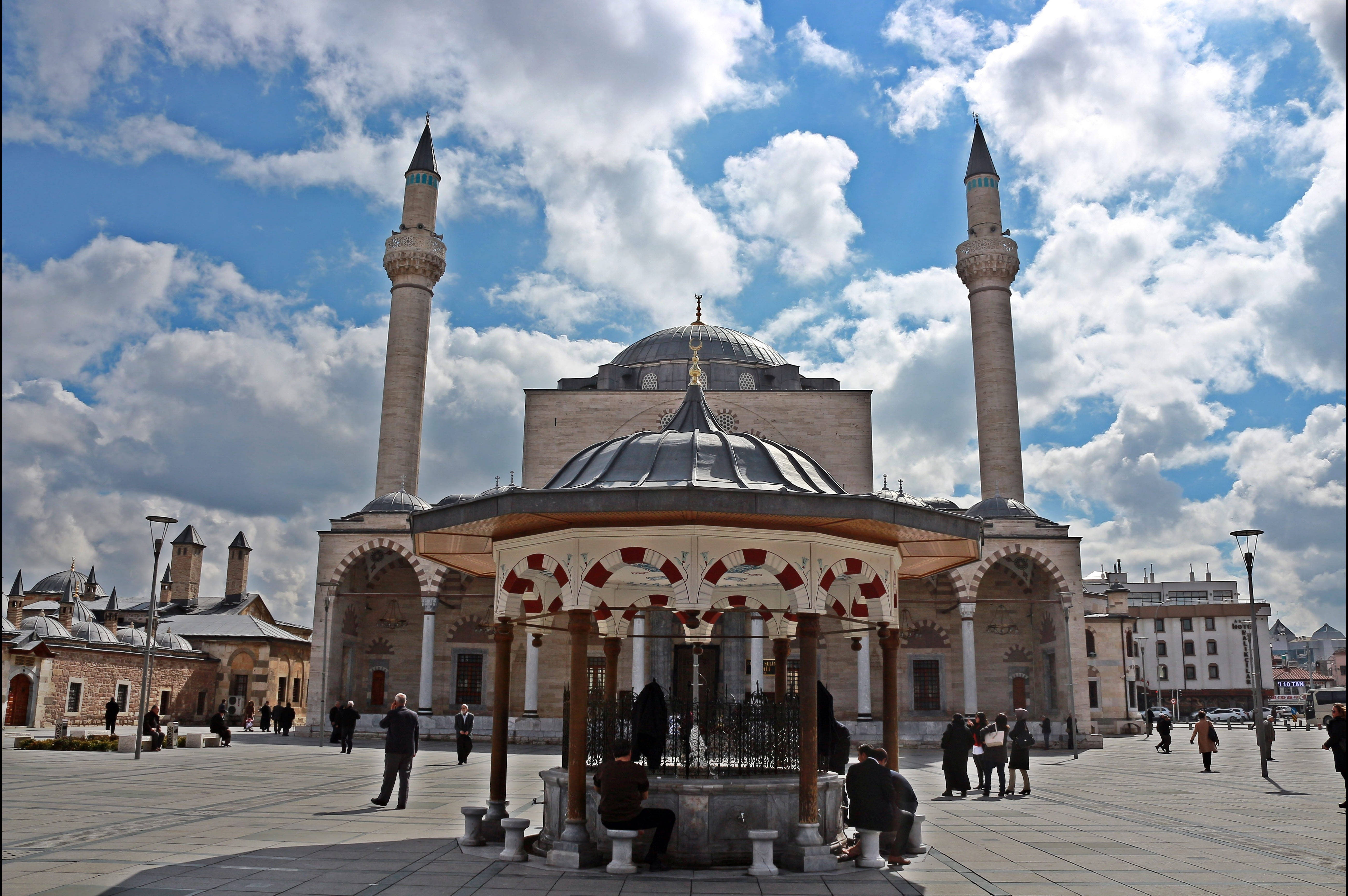 Konya 桌布 壁紙 — 一座宏偉的清真寺，擁有兩座高聳的宣禮塔和一個中央圓頂，矗立在明亮的藍天下，飄著蓬鬆的白雲，周圍是寬敞的廣場，有人們和一個裝飾性噴泉。