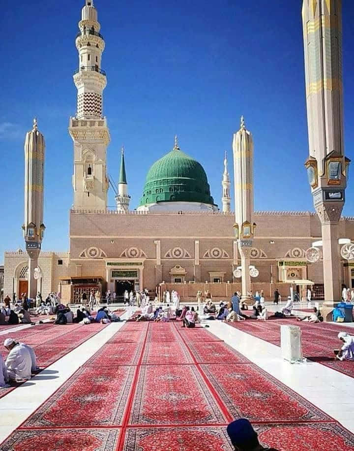 Medina 桌布 壁紙 — 一座宏偉的清真寺，擁有綠色圓頂和高聳的宣禮塔，在清澈的藍天下，周圍的信徒跪在紅色祈禱毯上。