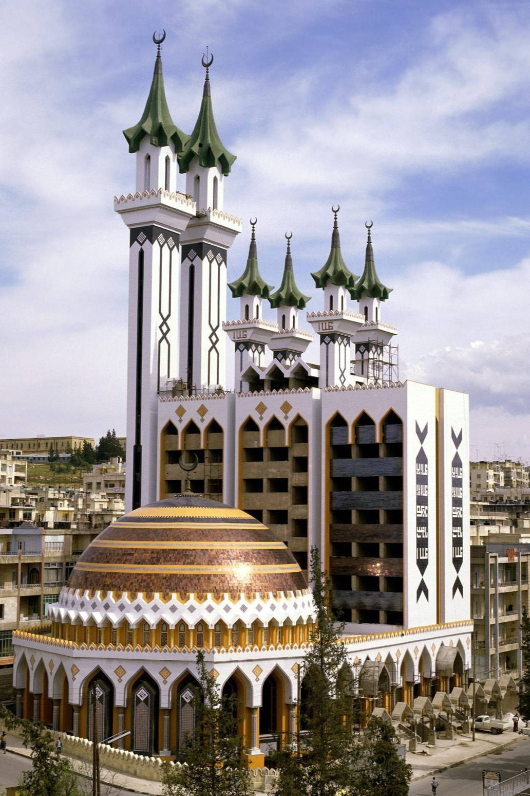Iraq 桌布 壁紙 — 一座宏偉的清真寺，擁有綠色尖塔和金色圓頂，在藍天映襯下矗立於城市環境中。