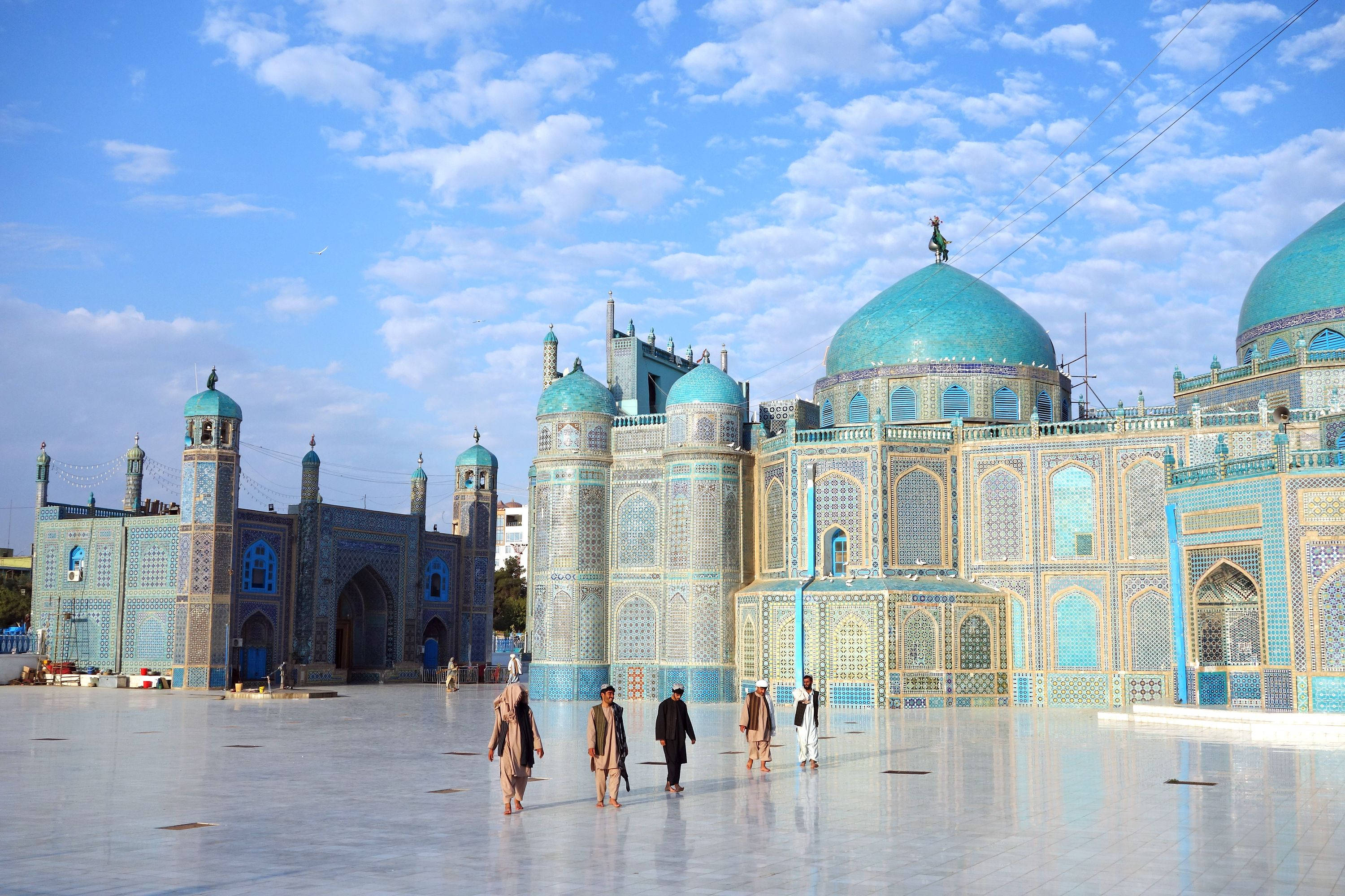 Afghanistan 桌布 壁紙 — 一座宏偉的清真寺，擁有藍綠色圓頂和精緻的瓷磚工藝，矗立在明亮的藍天和散落的雲朵下，人們走過其反射光線的庭院。