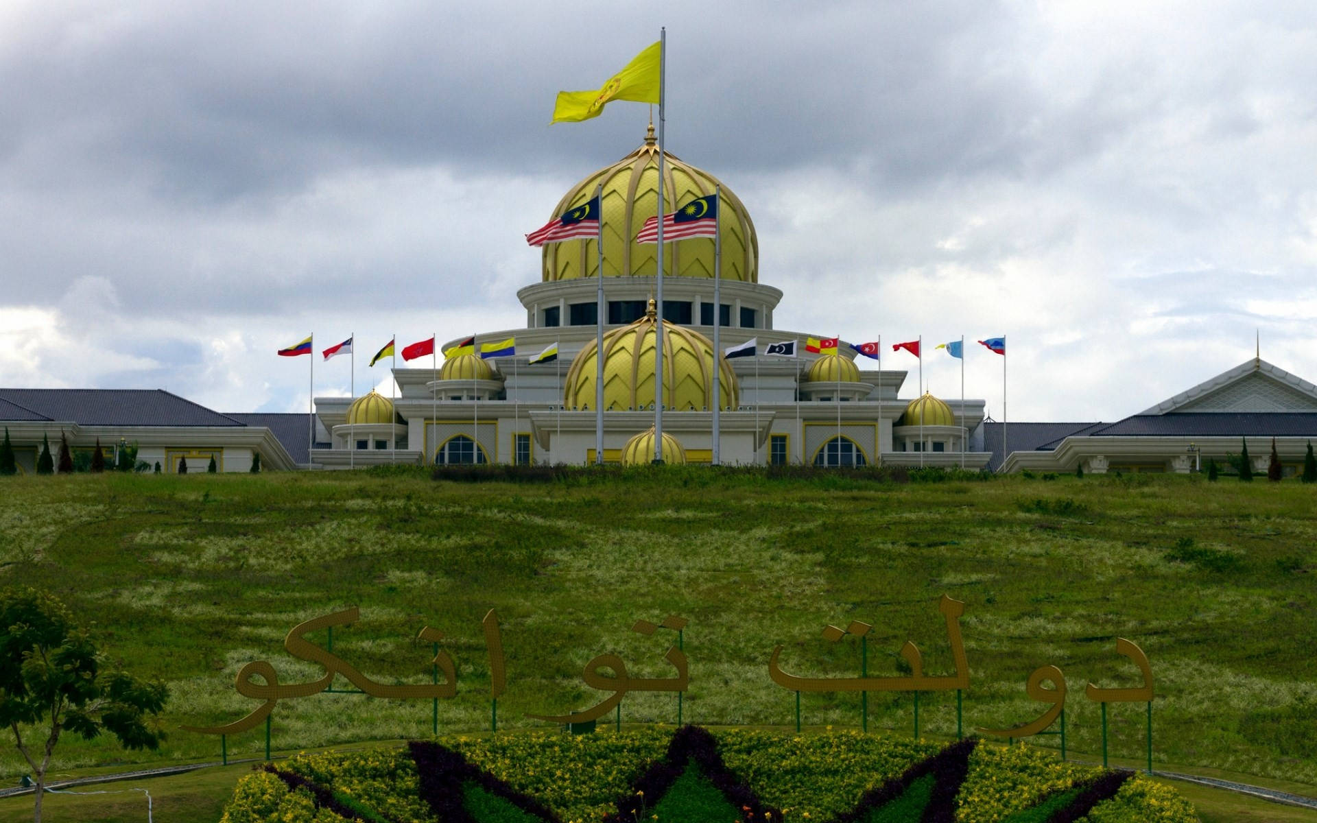 Jakarta 桌布 壁紙 — 一座宏偉的清真寺，擁有金色圓頂和馬來西亞國旗，矗立在雲天下鬱鬱蔥蔥的綠色山丘上。