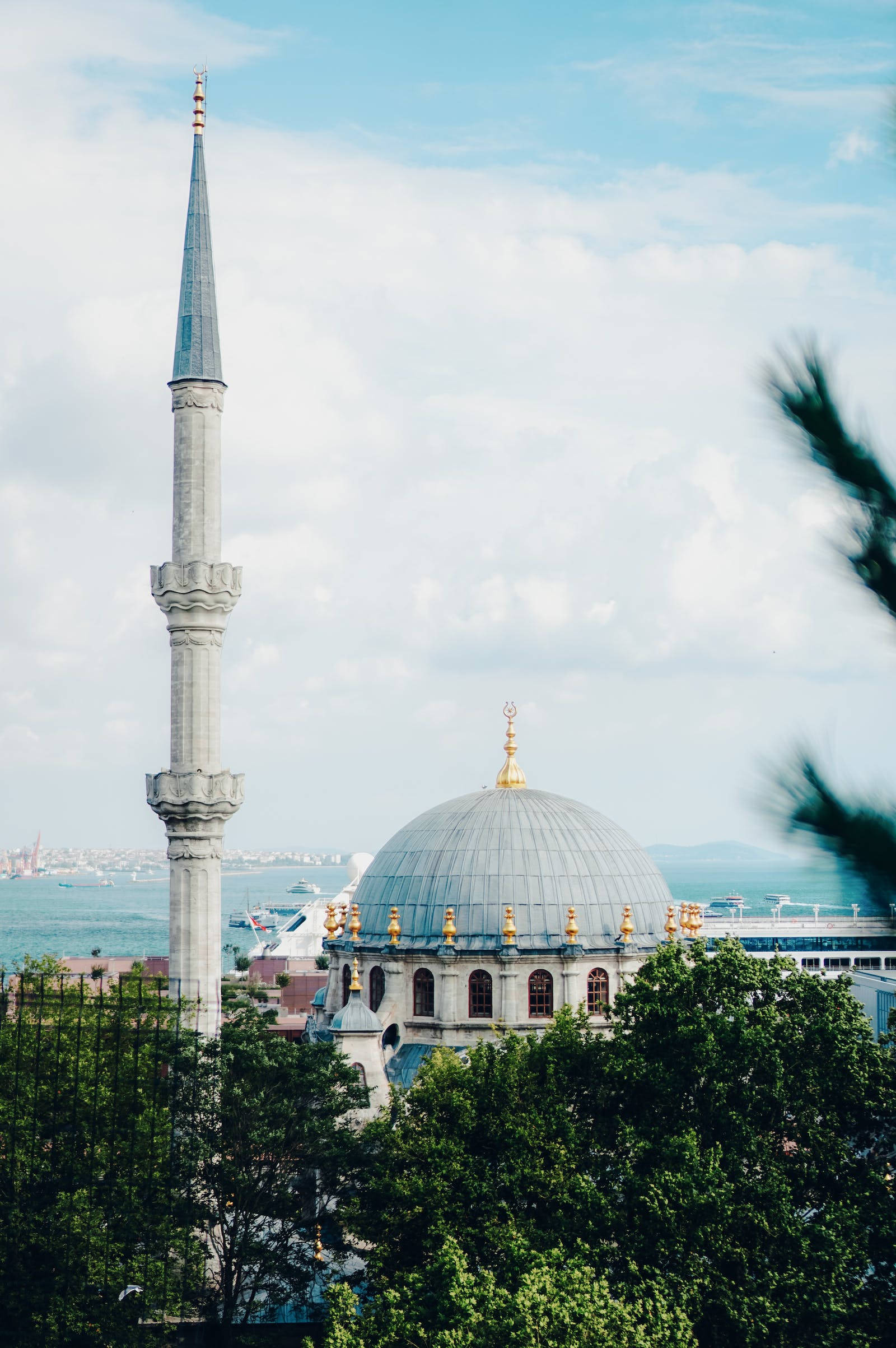HD Islamic 桌布 壁紙 — 一座宏偉的清真寺，擁有高聳的宣禮塔和圓頂屋頂，矗立在海濱旁，被茂密的綠樹環繞，天空部分多雲。