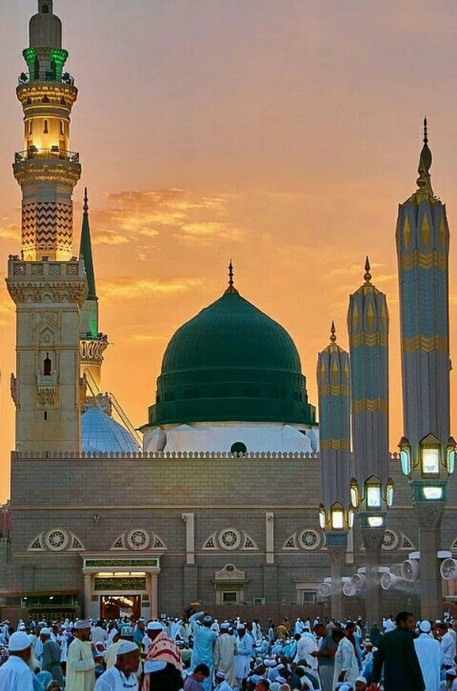 Medina 桌布 壁紙 — 一座宏偉的清真寺，綠色圓頂和宣禮塔沐浴在金色的日落光芒中，信徒們聚集在下方。