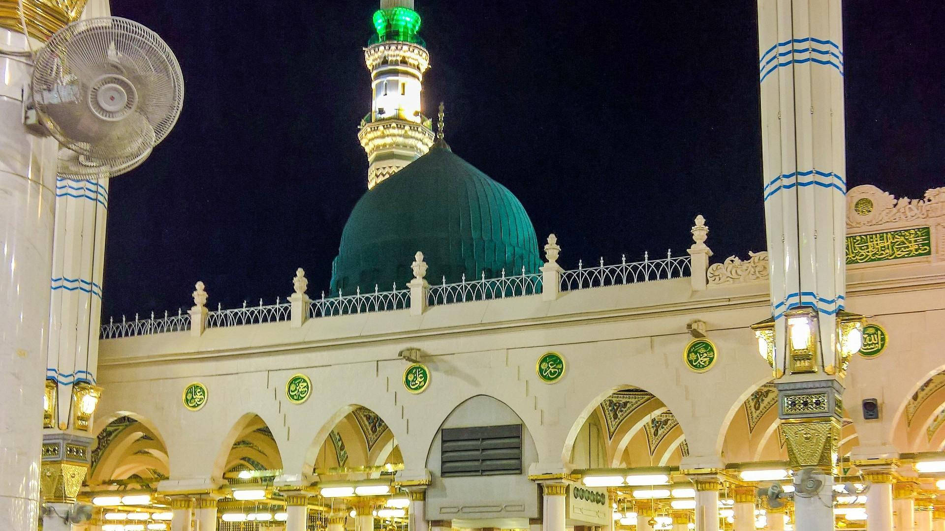 Madina 桌布 壁紙 — 一座宏偉的清真寺在夜晚被照亮，帶有發光的綠色圓頂和飾有藍色條紋的白色宣禮塔。