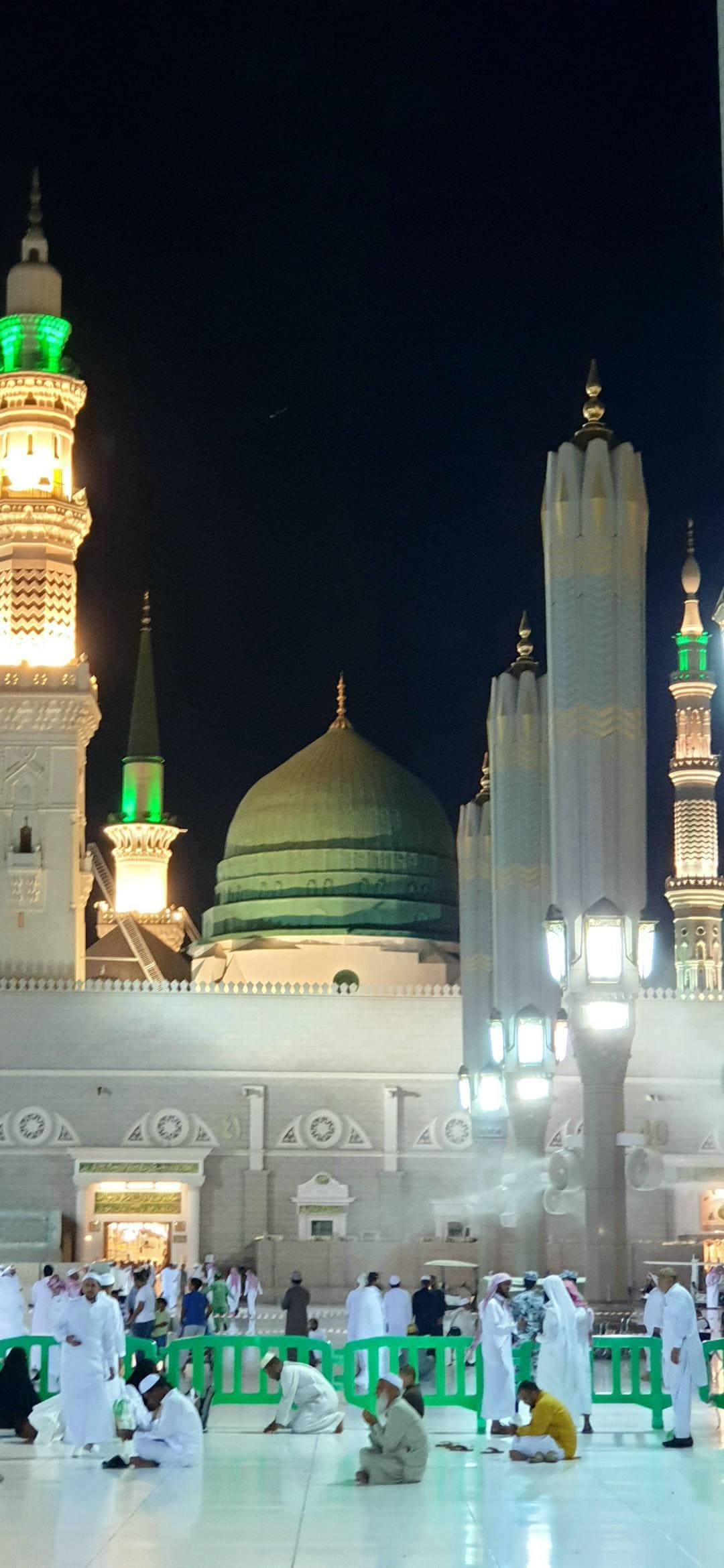 Madina 桌布 壁紙 — 一座宏偉的清真寺在夜晚被照亮，綠色圓頂和宣禮塔在黑暗的天空中閃耀，信徒們聚集祈禱。