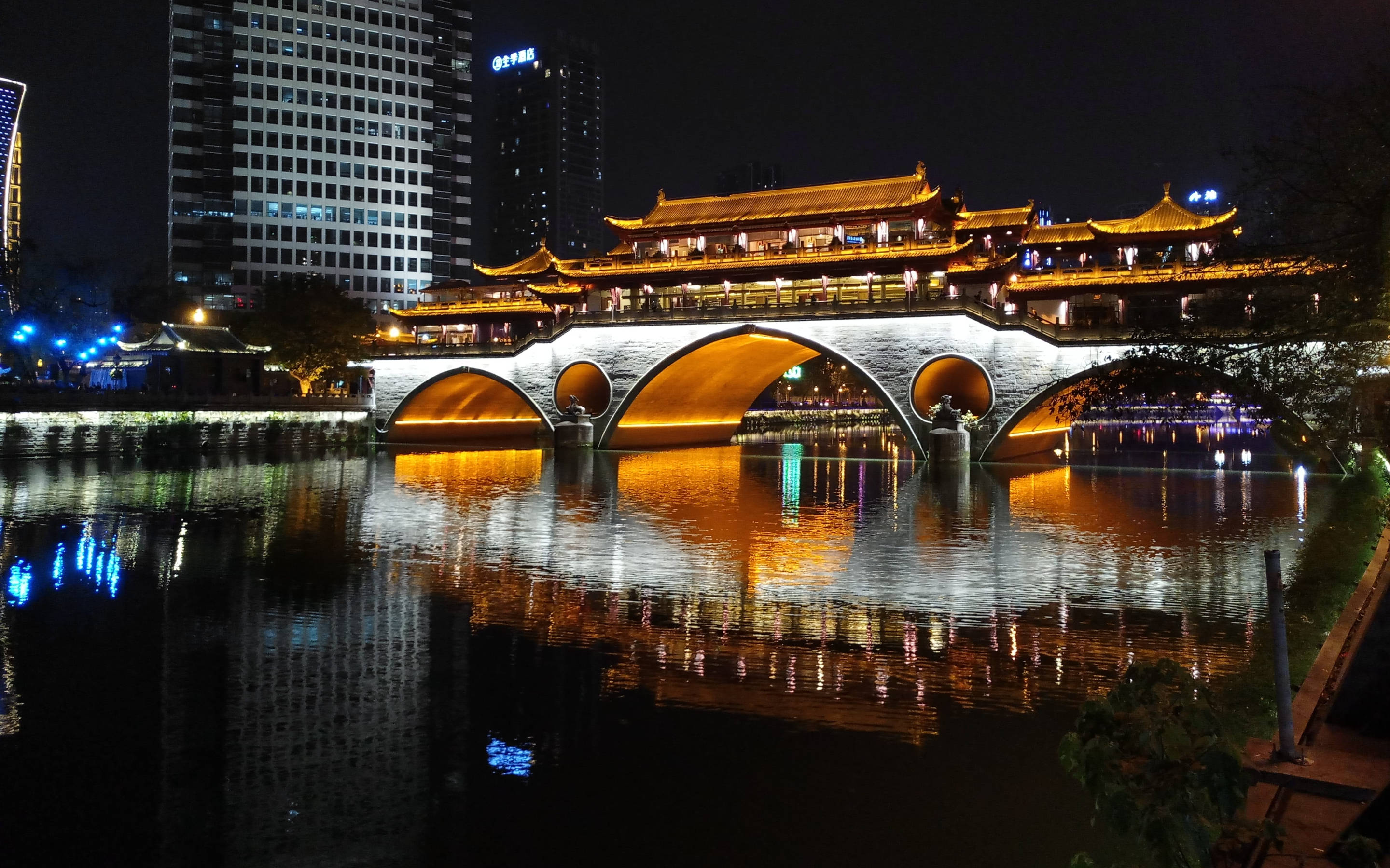 Chengdu 桌布 壁紙 — 一座宏偉的照明傳統中式橋樑，帶有拱形石質支撐，橫跨平靜的河流，反射金色燈光，背景是夜晚的現代摩天大樓。
