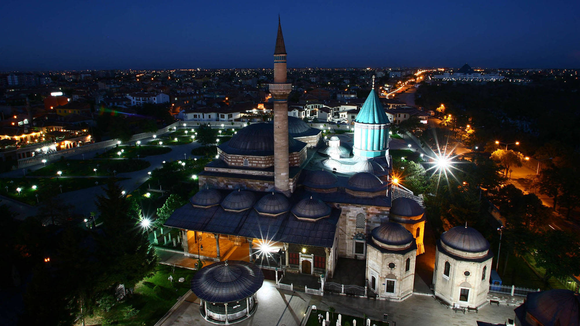 Konya 桌布 壁紙 — 一座宏偉的照明清真寺，擁有綠松石圓頂和高聳的宣禮塔，在夜晚莊嚴矗立，周圍環繞著閃爍的城市燈光和修剪整齊的花園。