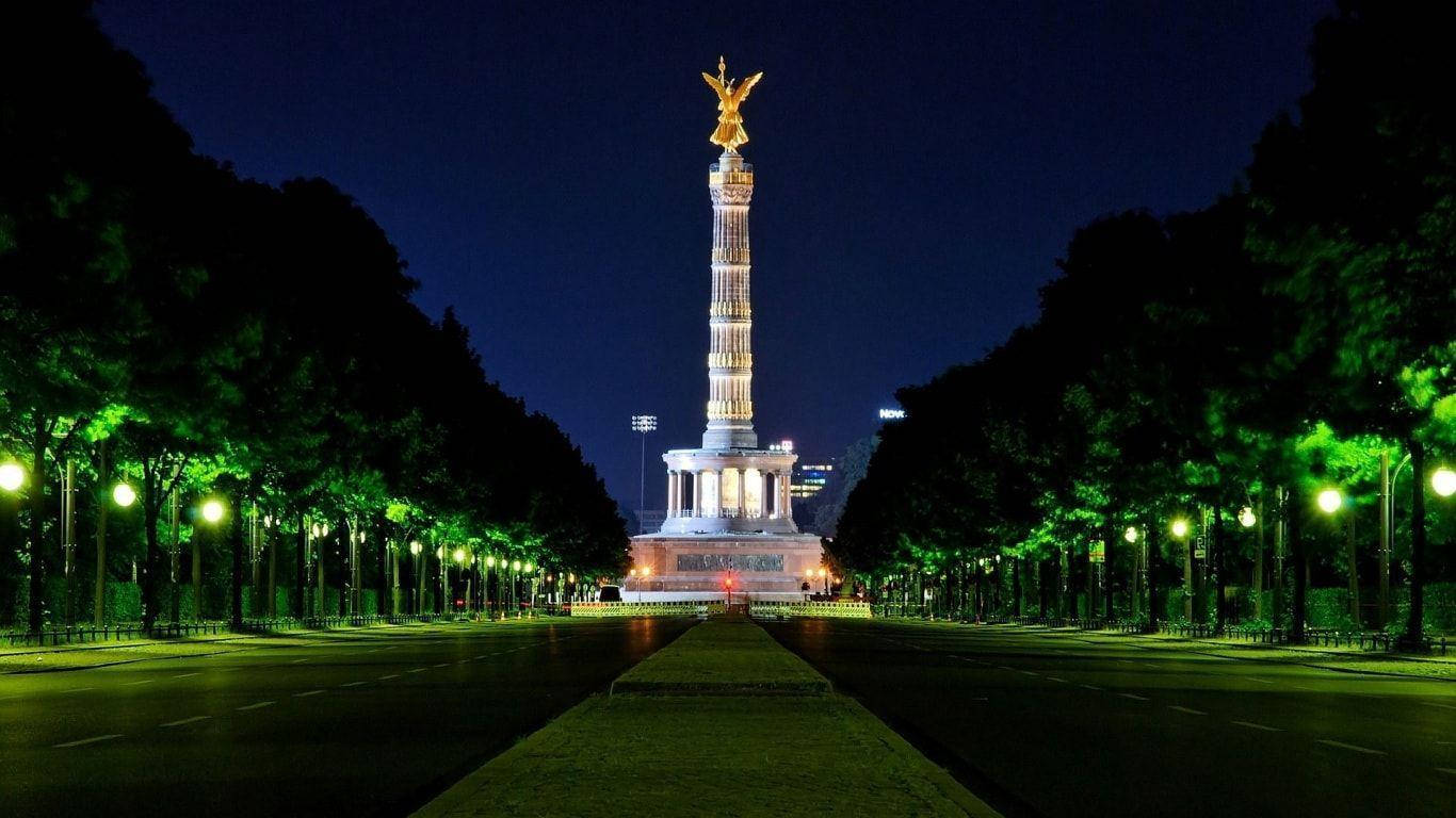 Berlin 桌布 壁紙 — 一座宏偉的照明紀念碑，頂部有金色天使，矗立在樹木成蔭的大道盡頭，在夜燈下閃耀。