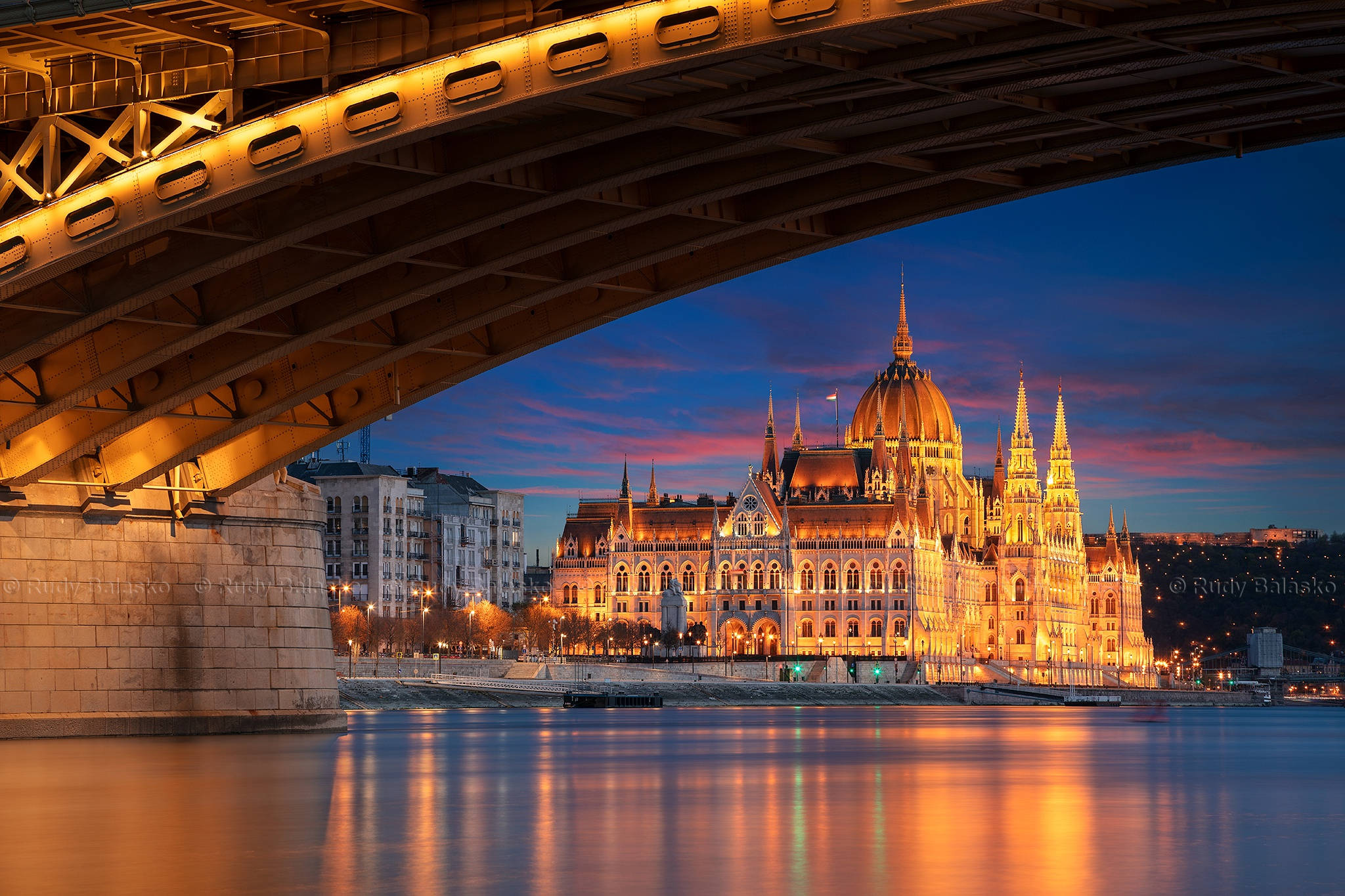 Hungary 桌布 壁紙 — 一座宏偉的照明議會大樓在黃昏時分於橋下閃耀金光，其倒影在平靜的河面上閃爍。