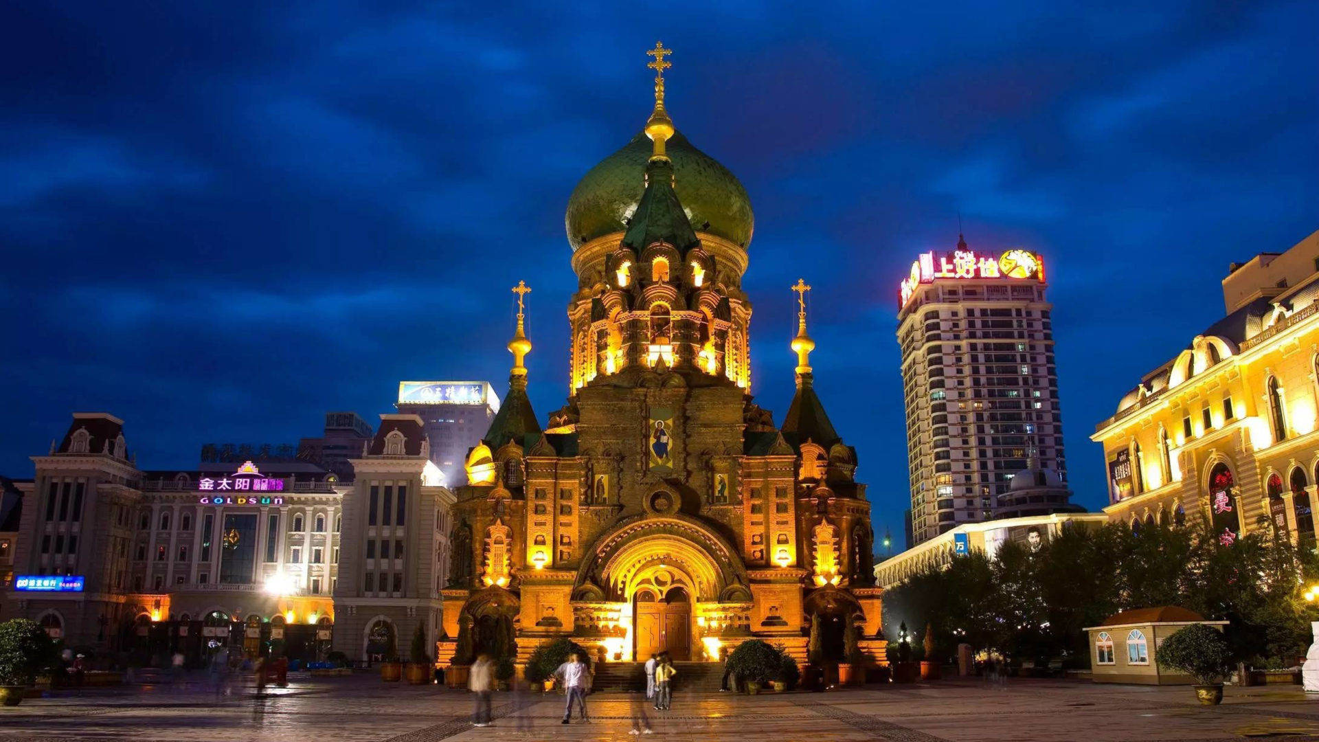 Harbin 桌布 壁紙 — 一座宏偉的燈光大教堂，擁有綠色圓頂和金色十字架，在黃昏時分雄偉地矗立在深藍色天空下，周圍環繞著現代城市建築和幾位行人。