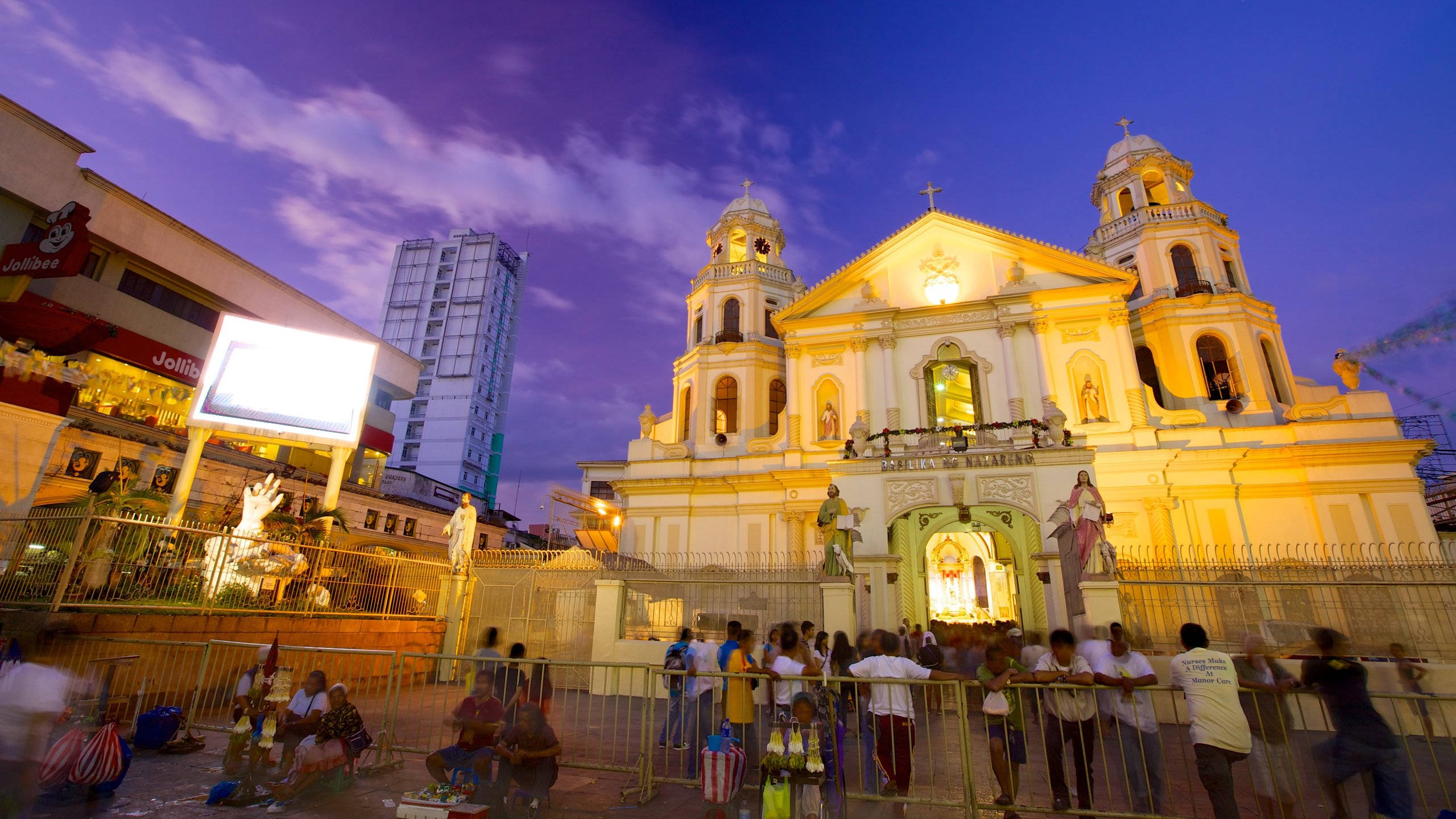 Manila 桌布 壁紙 — 一座宏偉的燈光教堂在暮色天空下溫暖地發光，人群聚集在其門外，身處繁華的城市環境中。