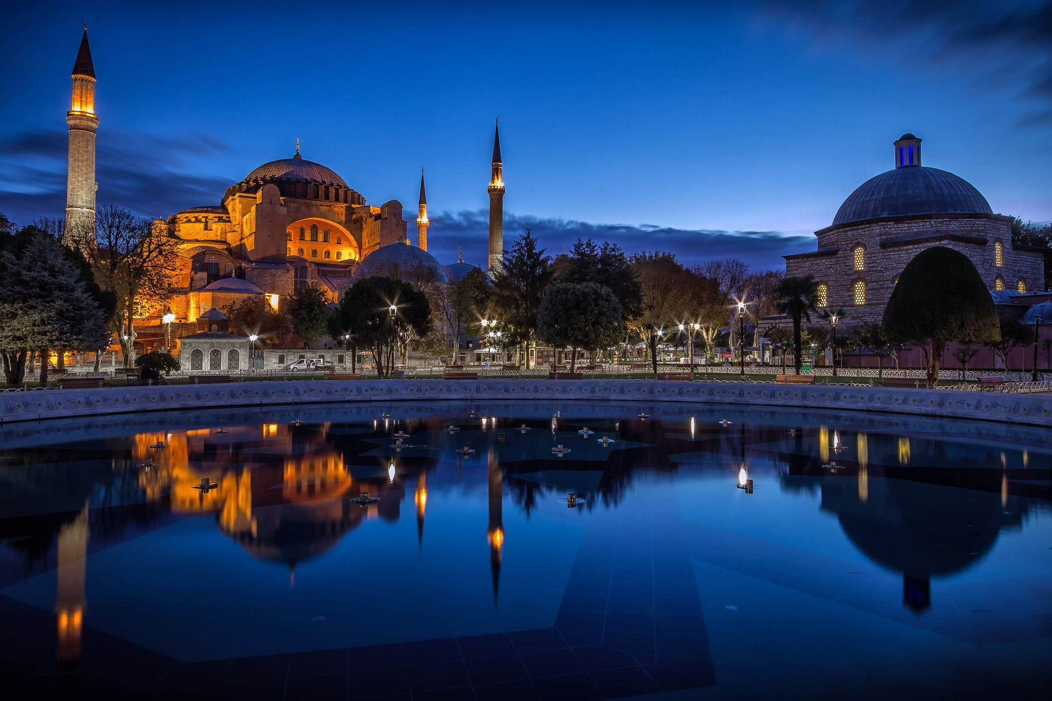 Istanbul 桌布 壁紙 — 一座宏偉的燈光清真寺，擁有圓頂和宣禮塔，在深藍色的暮色天空下完美地倒映在寧靜的池塘中。