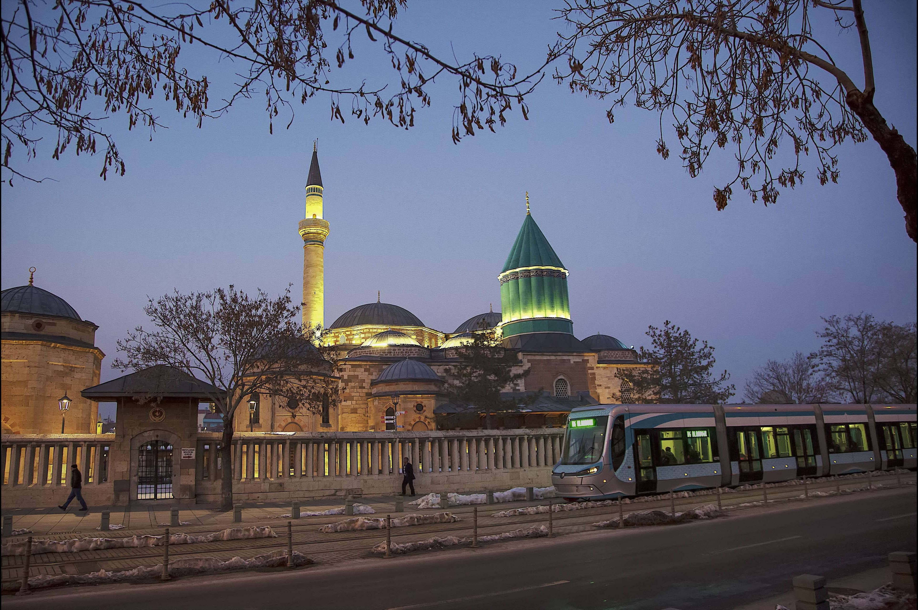 Konya 桌布 壁紙 — 一座宏偉的燈光清真寺，擁有綠色圓頂和宣禮塔，在黃昏時分矗立，一輛現代電車經過光禿的冬季樹木。