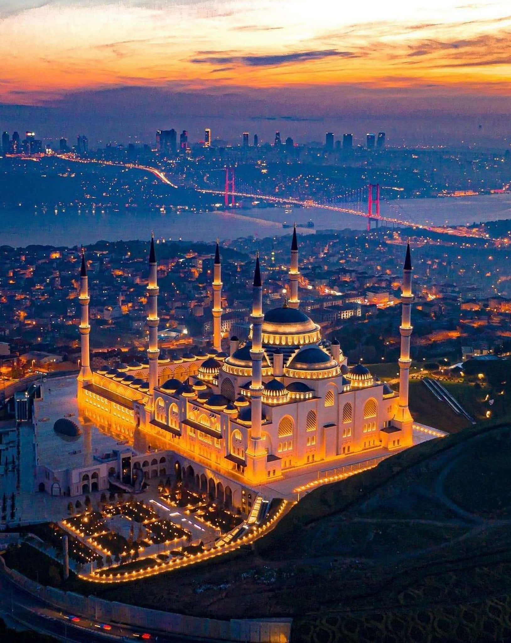 土耳其 桌布 壁紙 — 一座宏偉的燈光清真寺，有多個宣禮塔，俯瞰著日落時分廣闊的城市景觀，一座燈光橋樑橫跨水道。