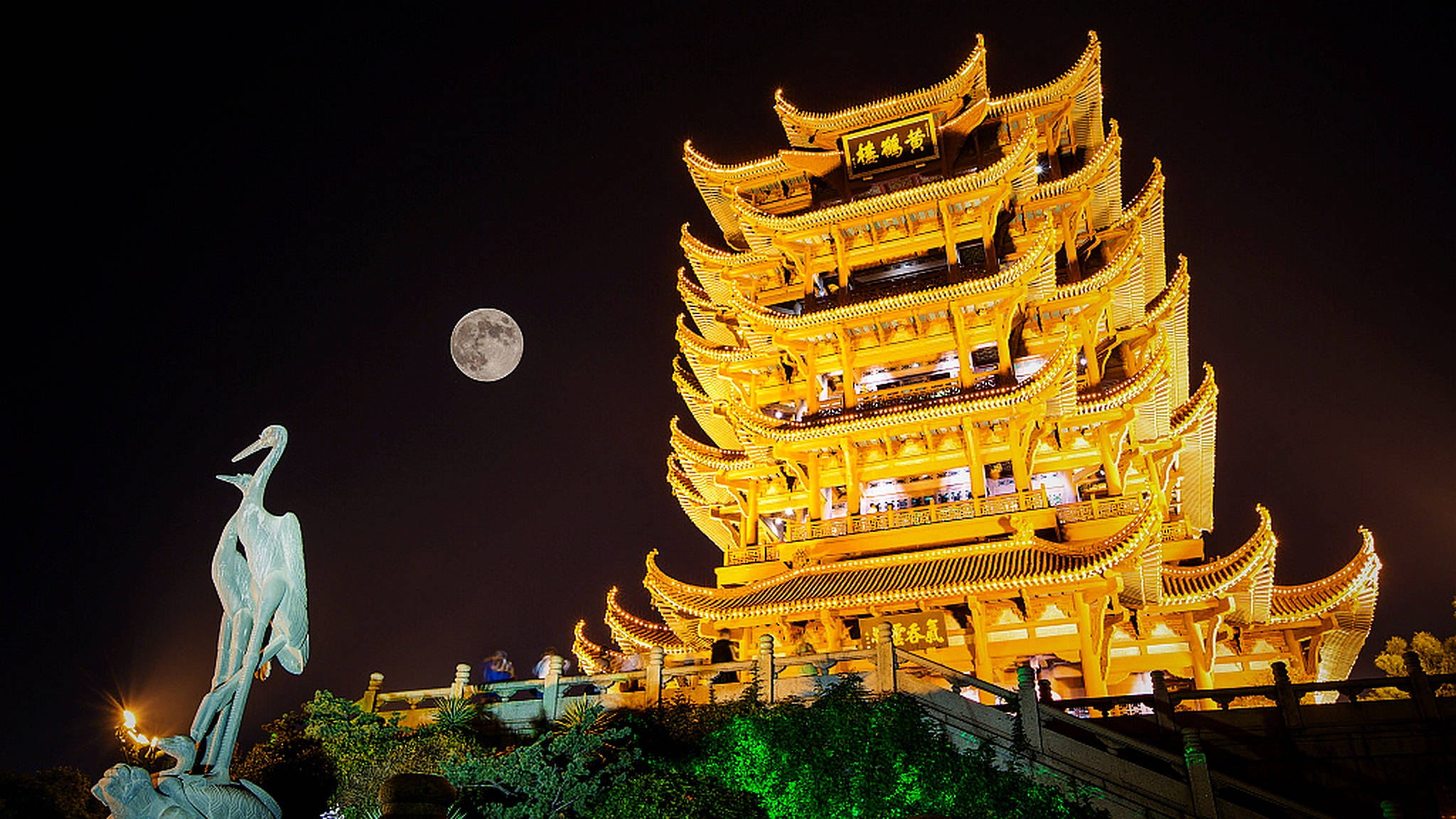 Wuhan 桌布 壁紙 — 一座宏偉的燈光璀璨的中國寶塔在滿月下閃耀金光，旁邊有一座鶴雕像，夜晚時分。