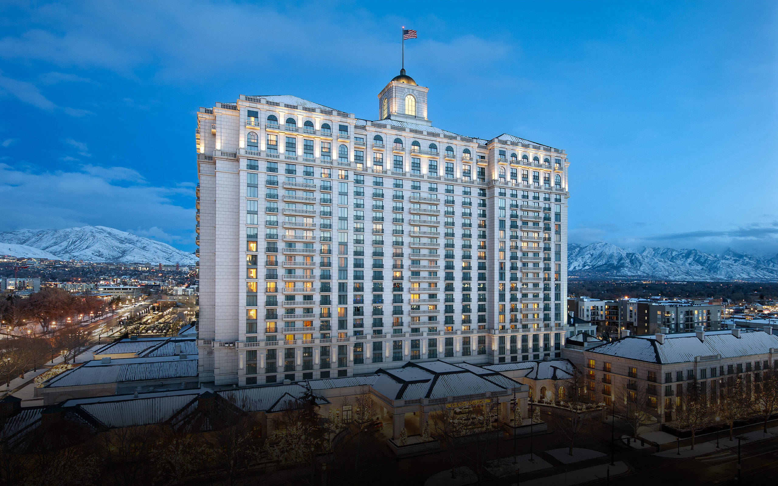 Salt Lake City 桌布 壁紙 — 一座宏偉的燈光酒店聳立在雪景城市景觀之上，雄偉的雪山在暮色天空下閃耀。