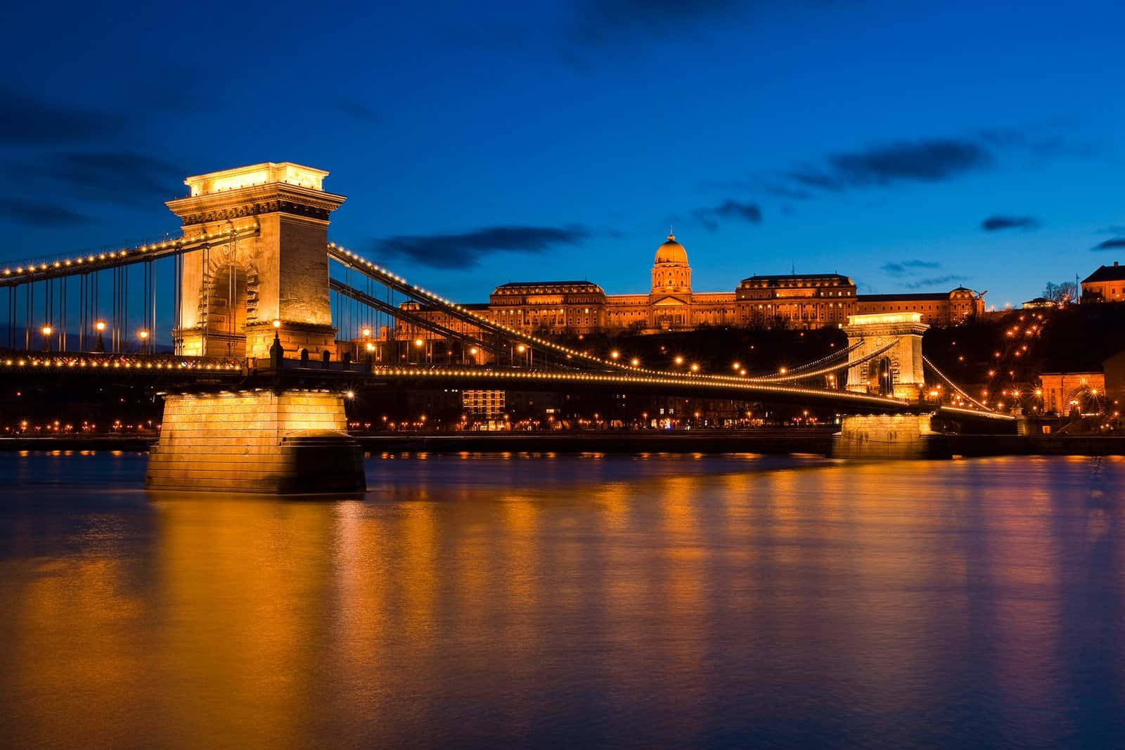 budapest 桌布 壁紙 — 一座宏偉的燈光鏈橋橫跨在暮色中平靜的河面上，歷史建築在深藍色天空的映襯下溫暖地發光。