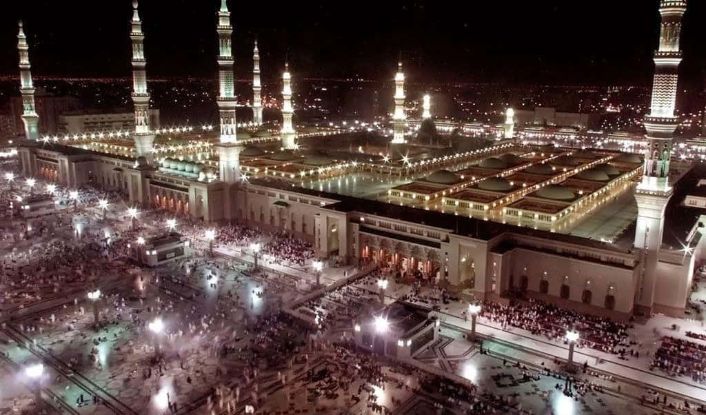 Medina 桌布 壁紙 — 一座宏偉的燈火通明的清真寺的夜間鳥瞰圖，高聳的宣禮塔和廣場上聚集的大量人群。