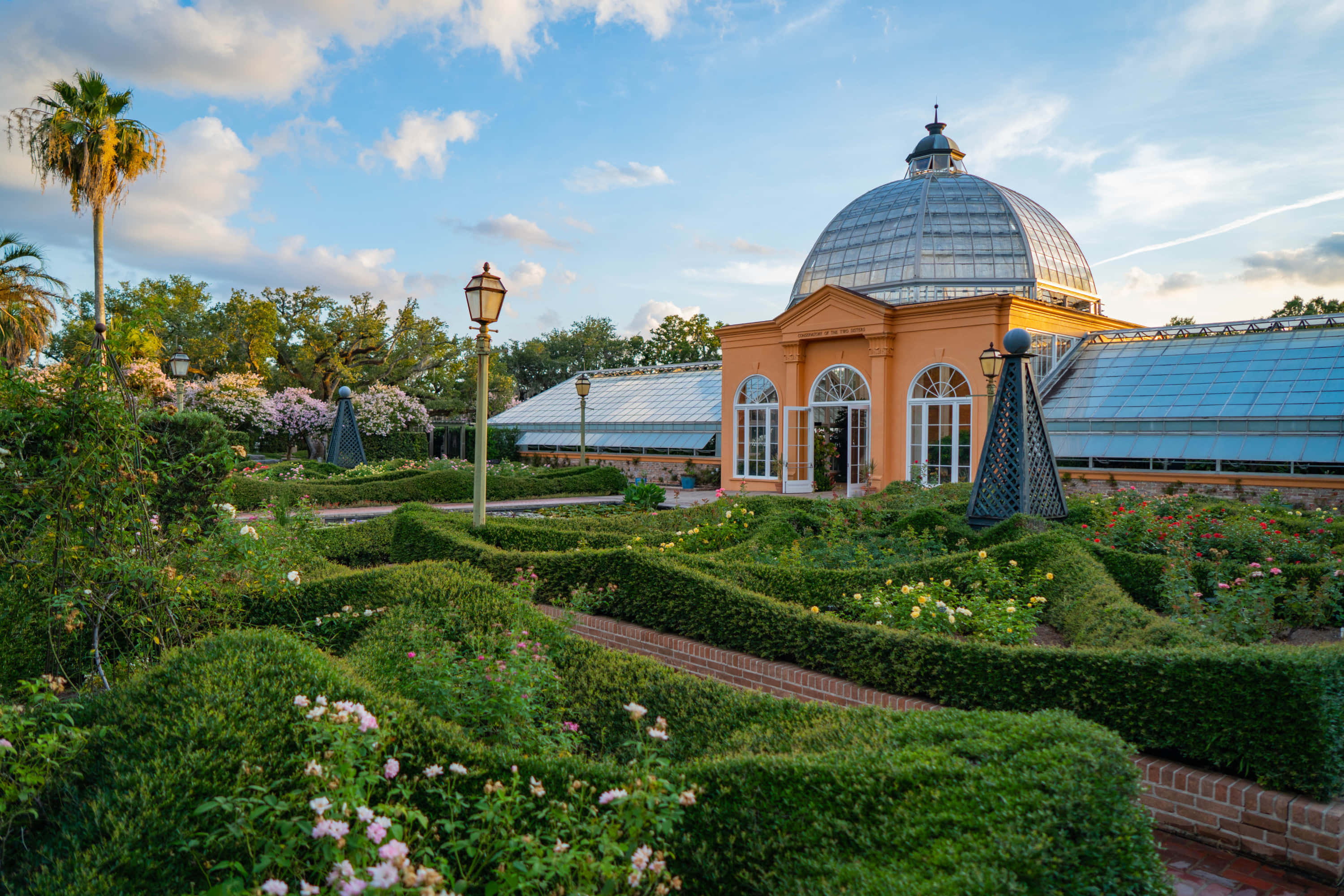 Botanical Garden 桌布 壁紙 — 一座宏偉的玻璃圓頂溫室，坐落在精心修剪的玫瑰園中，上方是藍天白雲。