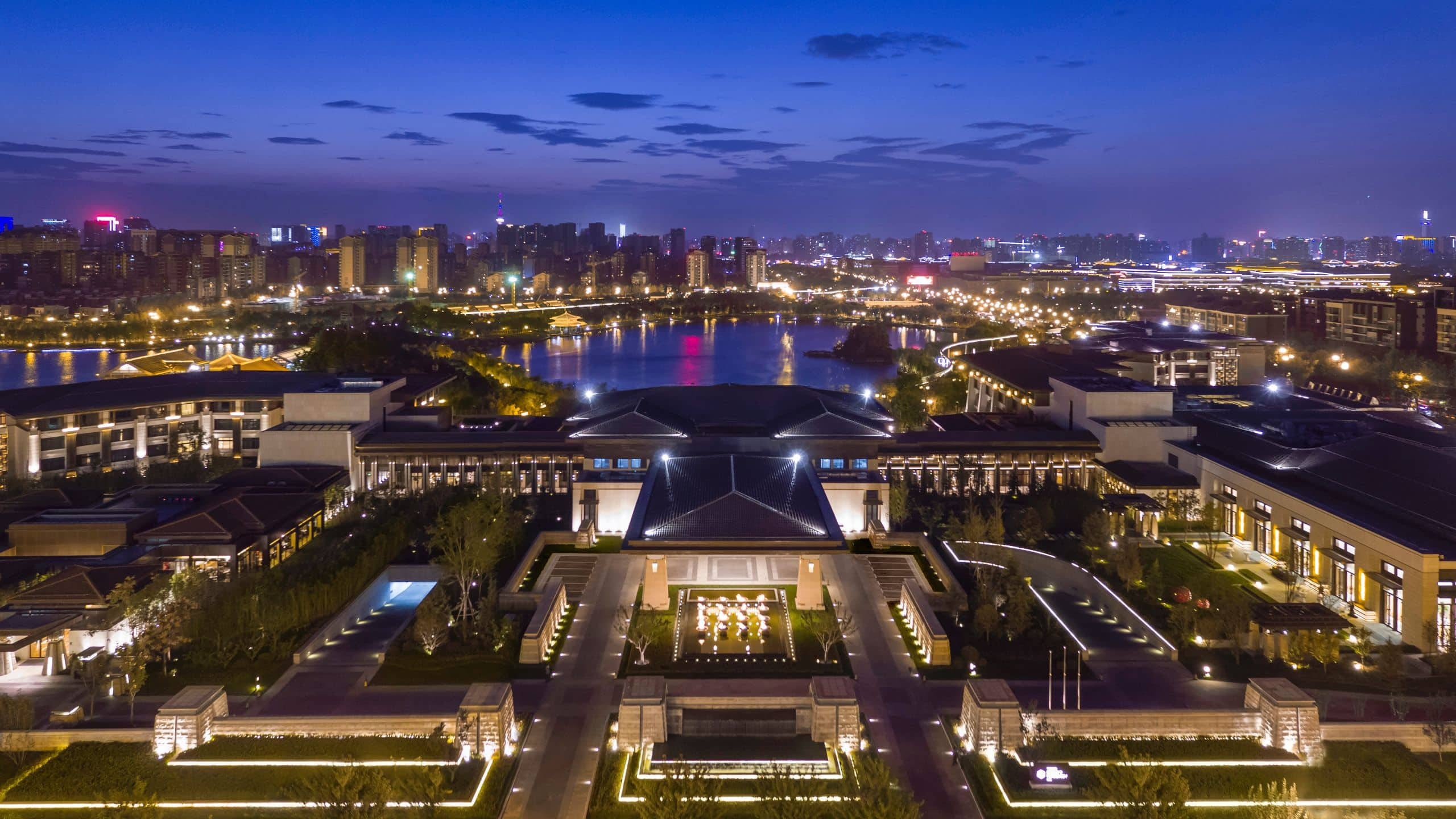 Xian 桌布 壁紙 — 一座宏偉的現代建築群在黃昏時分被溫暖的燈光照亮，燈光倒映在平靜的河面上，周圍是廣闊的城市景觀。