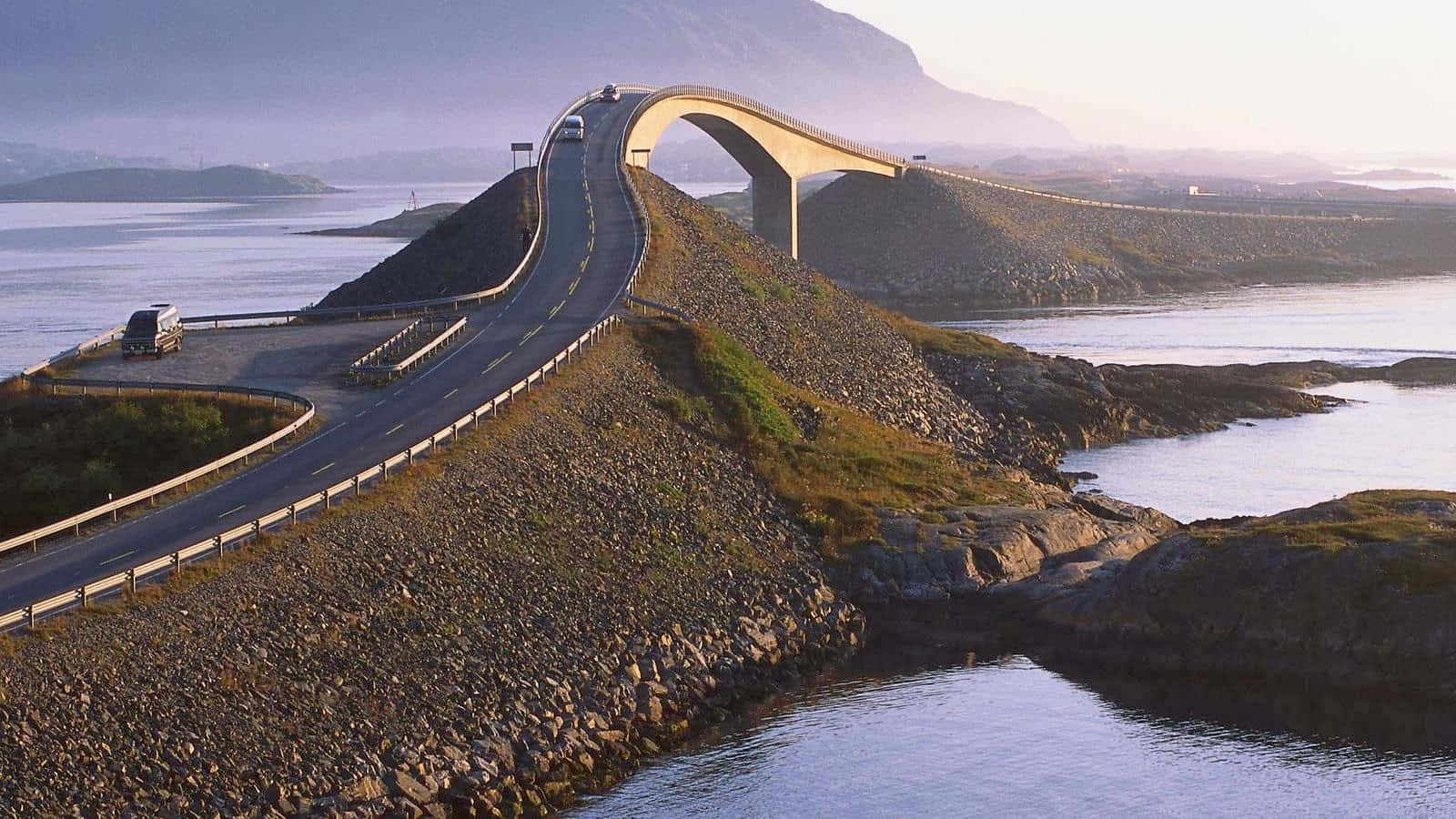 Road 桌布 壁紙 — 一座宏偉的現代橋樑優雅地拱起在岩石峽灣之上，汽車在黃金時段沿著蜿蜒的道路行駛。
