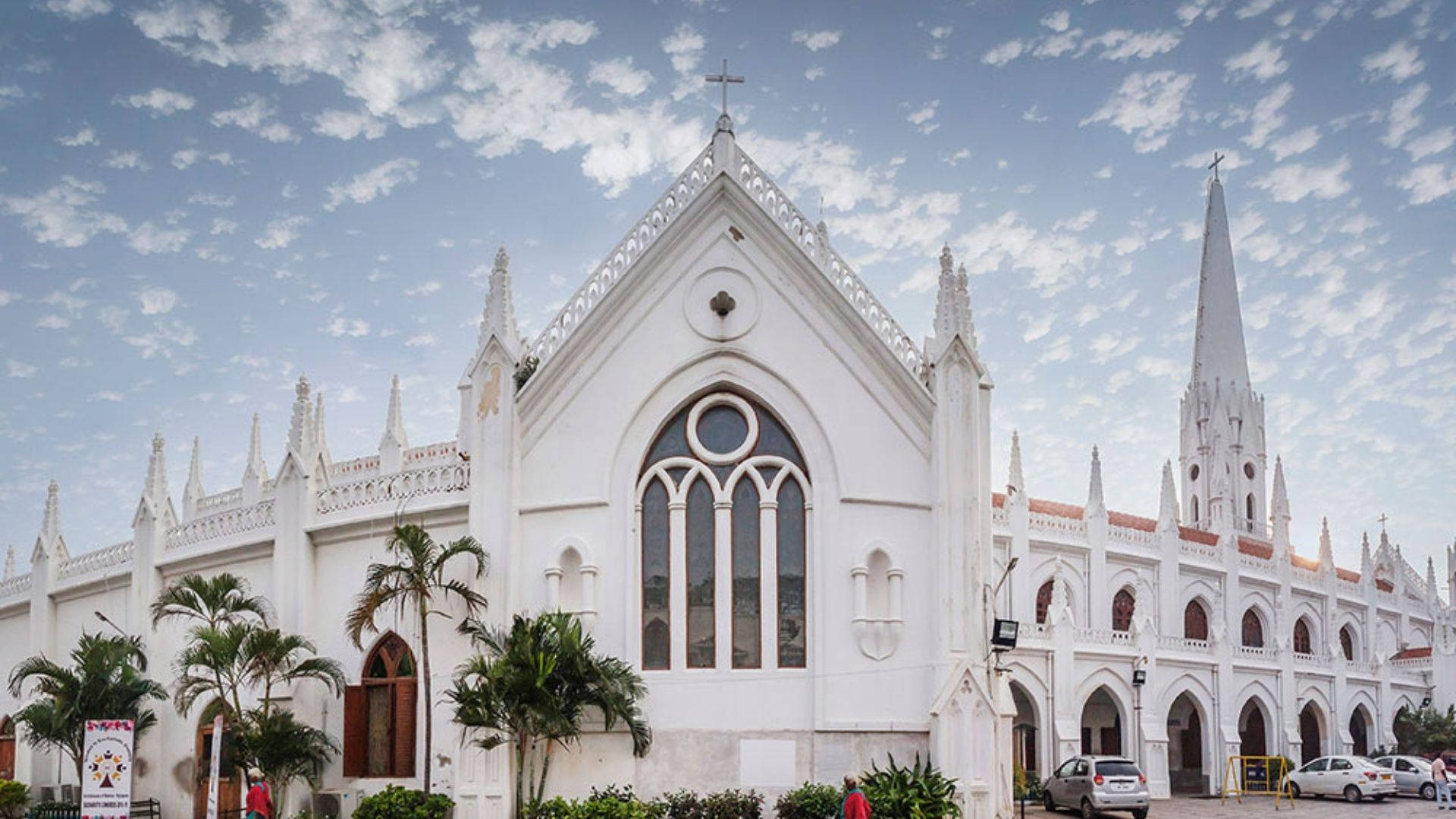 Chennai 桌布 壁紙 — 一座宏偉的白色哥德式教堂，擁有高聳的尖頂和拱形窗戶，矗立在部分多雲的天空下，附近有棕櫚樹。