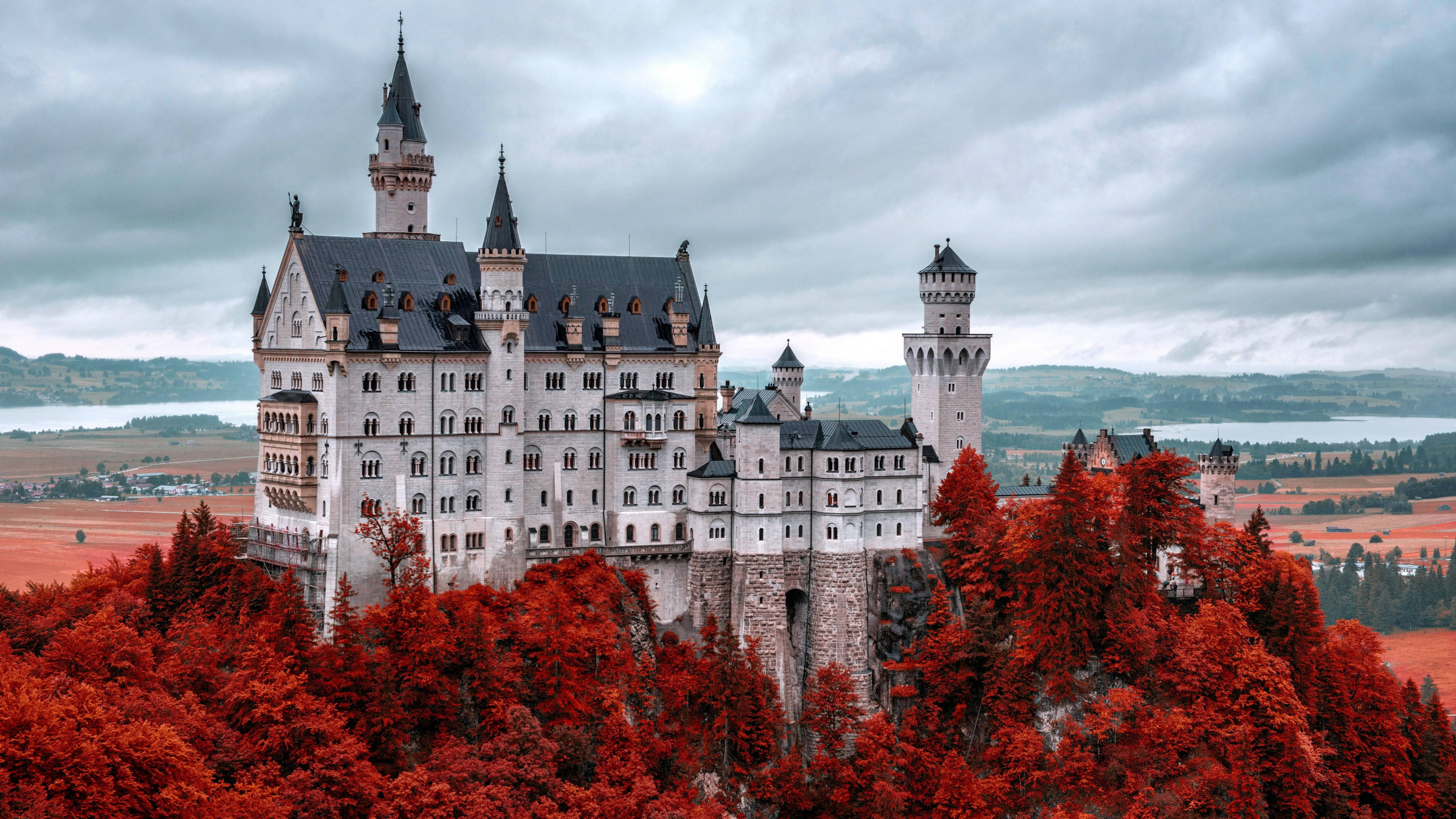 Neuschwanstein Castle 桌布 壁紙 — 一座宏偉的白色城堡，帶有黑暗的尖頂，聳立在佈滿火紅秋樹的山丘上，天空陰沉灰暗。