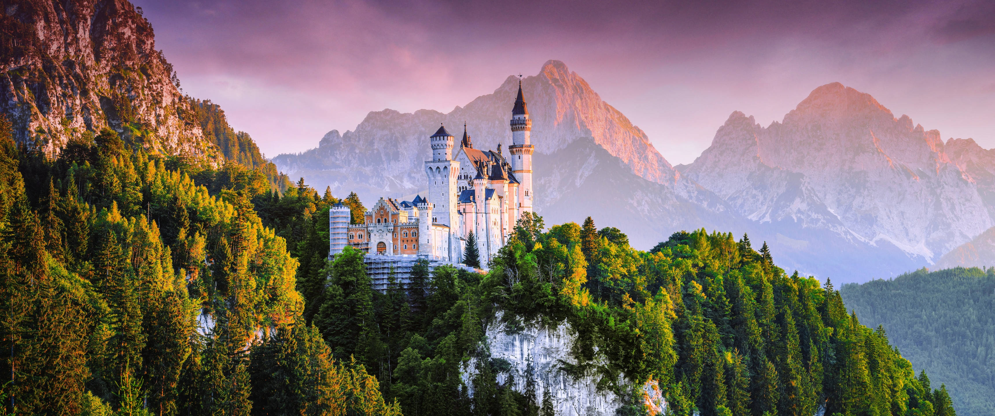 Neuschwanstein Castle 桌布 壁紙 — 一座宏偉的白色城堡坐落在樹木繁茂的山丘上，白雪覆蓋的山峰在紫色日落天空下閃耀。