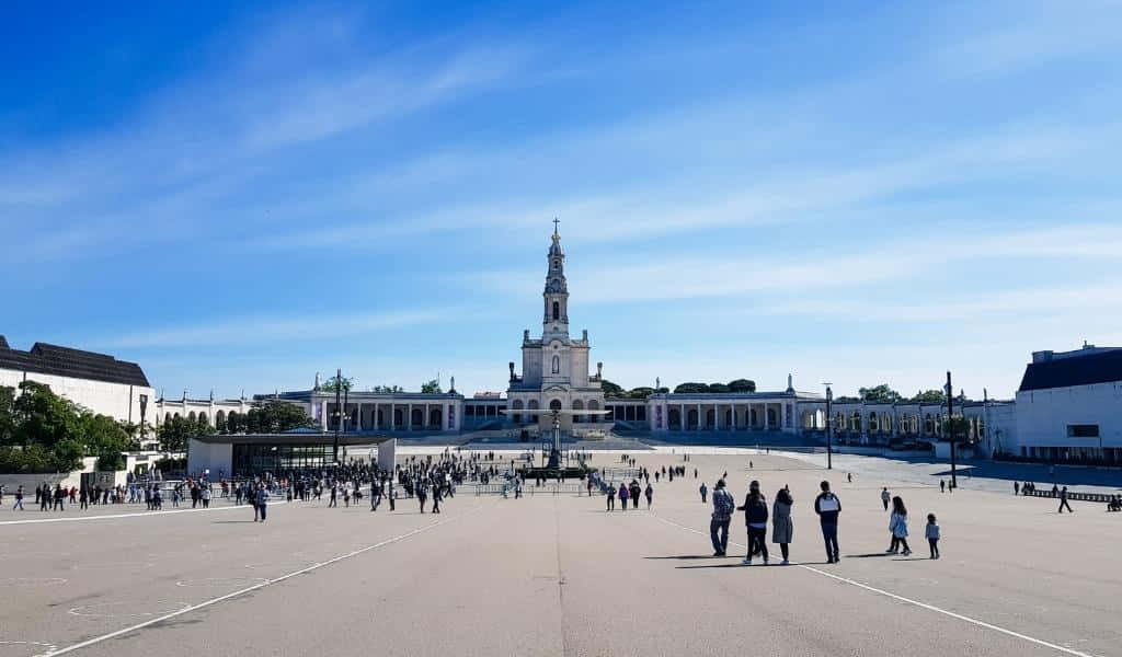 Portugal 桌布 壁紙 — 一座宏偉的白色大教堂，帶有高聳的尖塔，矗立在一個廣闊廣場的盡頭，廣場上擠滿了人，天空湛藍。