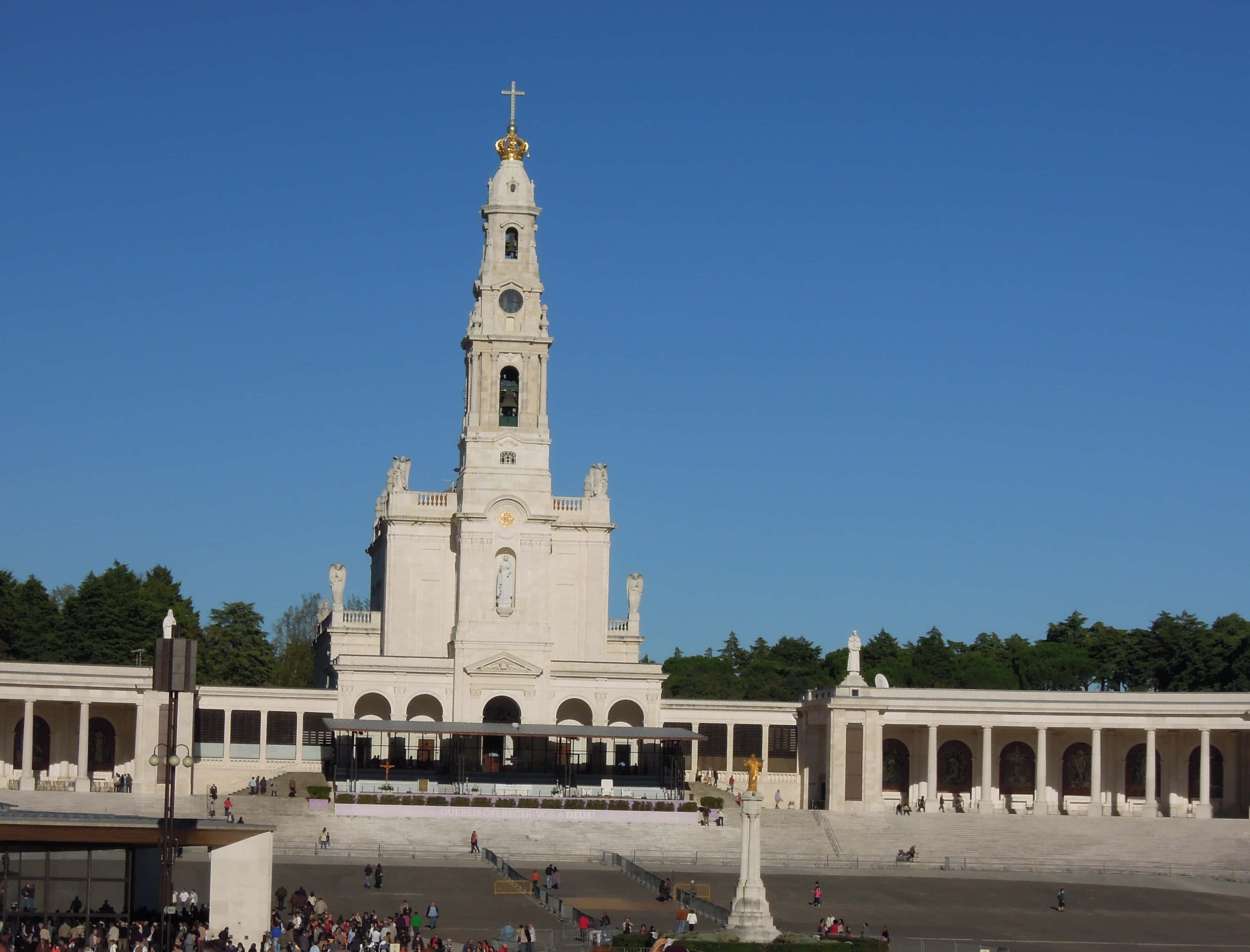 Portugal 桌布 壁紙 — 一座宏偉的白色大教堂，擁有高聳的鐘樓和金色十字架，矗立在清澈的藍天下，周圍環繞著人群和柱廊。