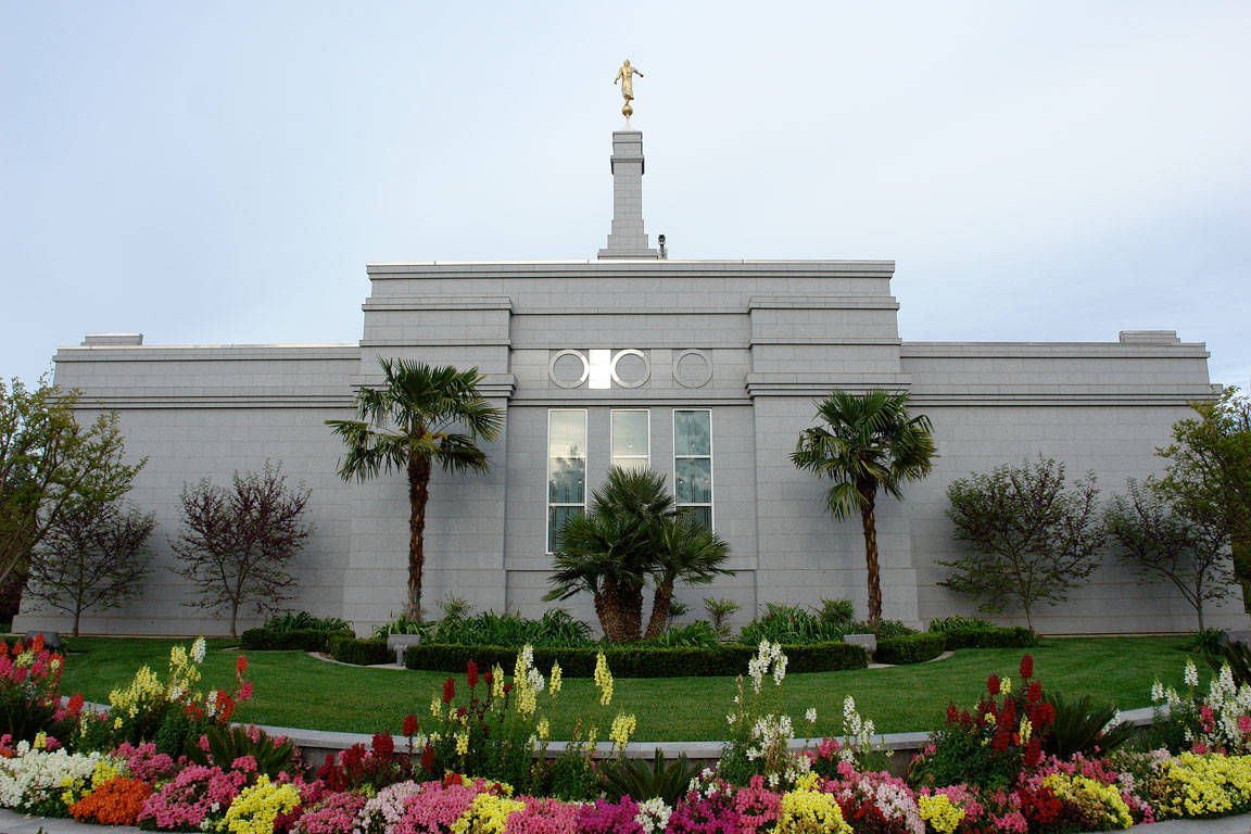Church 桌布 壁紙 — 一座宏偉的白色寺廟，頂部有金色雕像，矗立在色彩繽紛的花壇和棕櫚樹後方，天空蒼白。