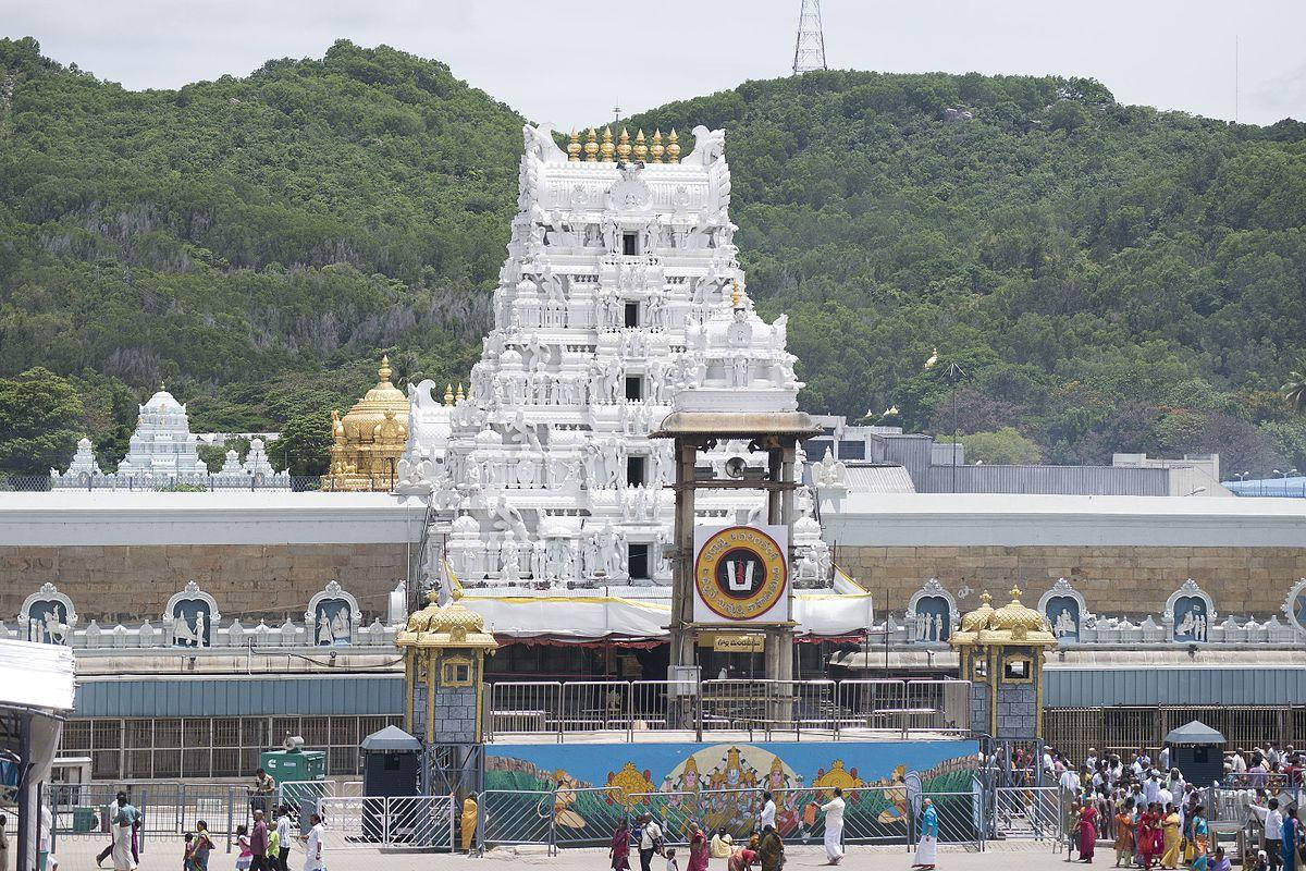 Venkateswara 桌布 壁紙 — 一座宏偉的白色寺廟帶有金色點綴，矗立在翠綠的山丘前，信徒們聚集在入口處。