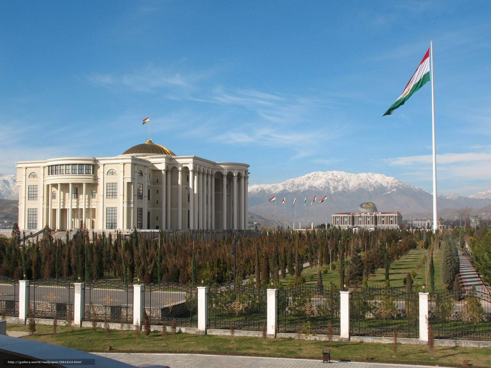 Tajikistan 桌布 壁紙 — 一座宏偉的白色政府建築，帶有金色圓頂，矗立在白雪覆蓋的山脈前，藍天白雲下，一面巨大的國旗飄揚。