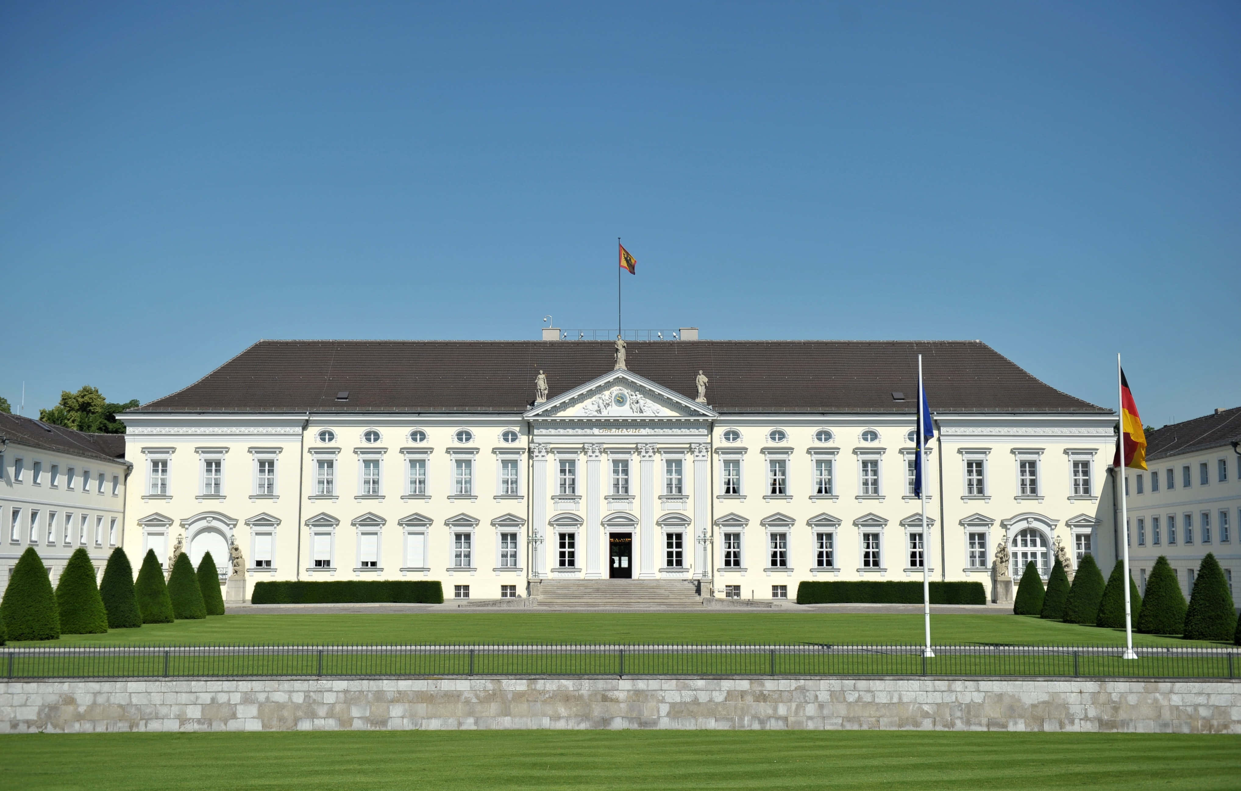 Berlin 桌布 壁紙 — 一座宏偉的白色新古典主義宮殿，帶有深色屋頂，矗立在清澈的藍天下，兩旁是修剪整齊的樹籬和德國國旗。
