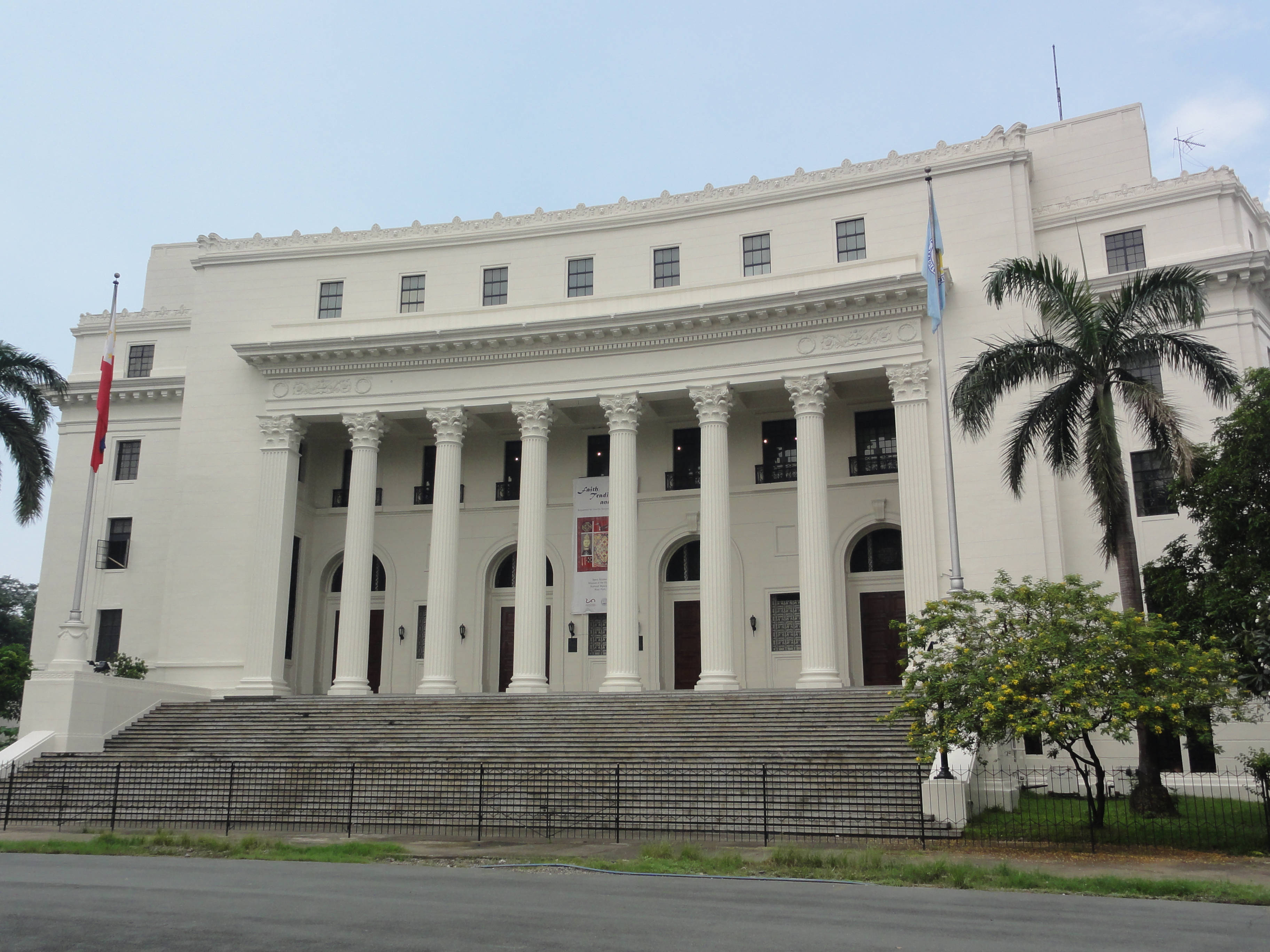 Manila 桌布 壁紙 — 一座宏偉的白色新古典主義建築，擁有高大的柱子和寬闊的台階，矗立在蒼白的天空下，兩旁種有棕櫚樹。