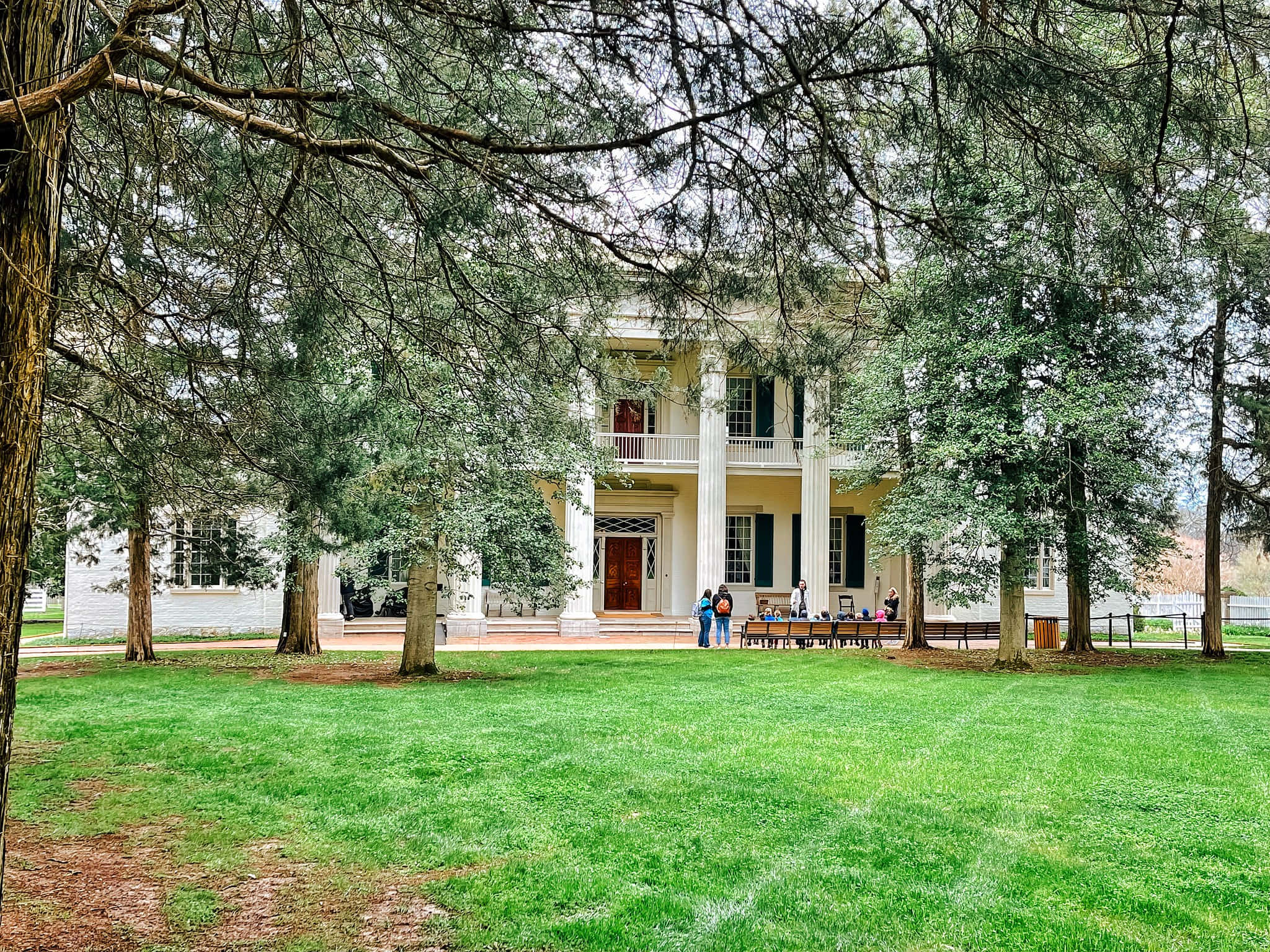 Mansion 桌布 壁紙 — 一座宏偉的白色柱式大宅，被高聳的常綠樹和充滿活力的綠色草坪環繞，訪客聚集在門廊附近。