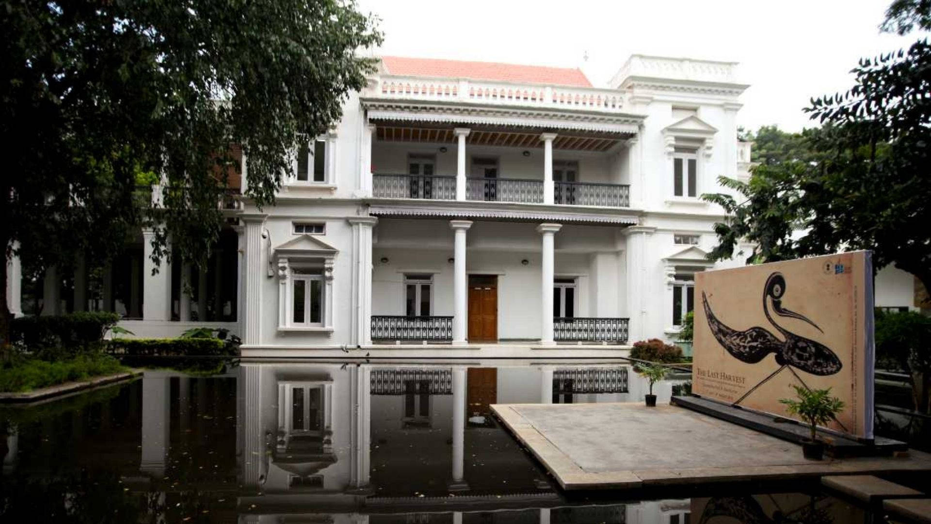 Bangalore 桌布 壁紙 — 一座宏偉的白色殖民風格大宅，帶有柱子和陽台，完美倒映在平靜的池塘中，周圍環繞著茂密的綠樹。