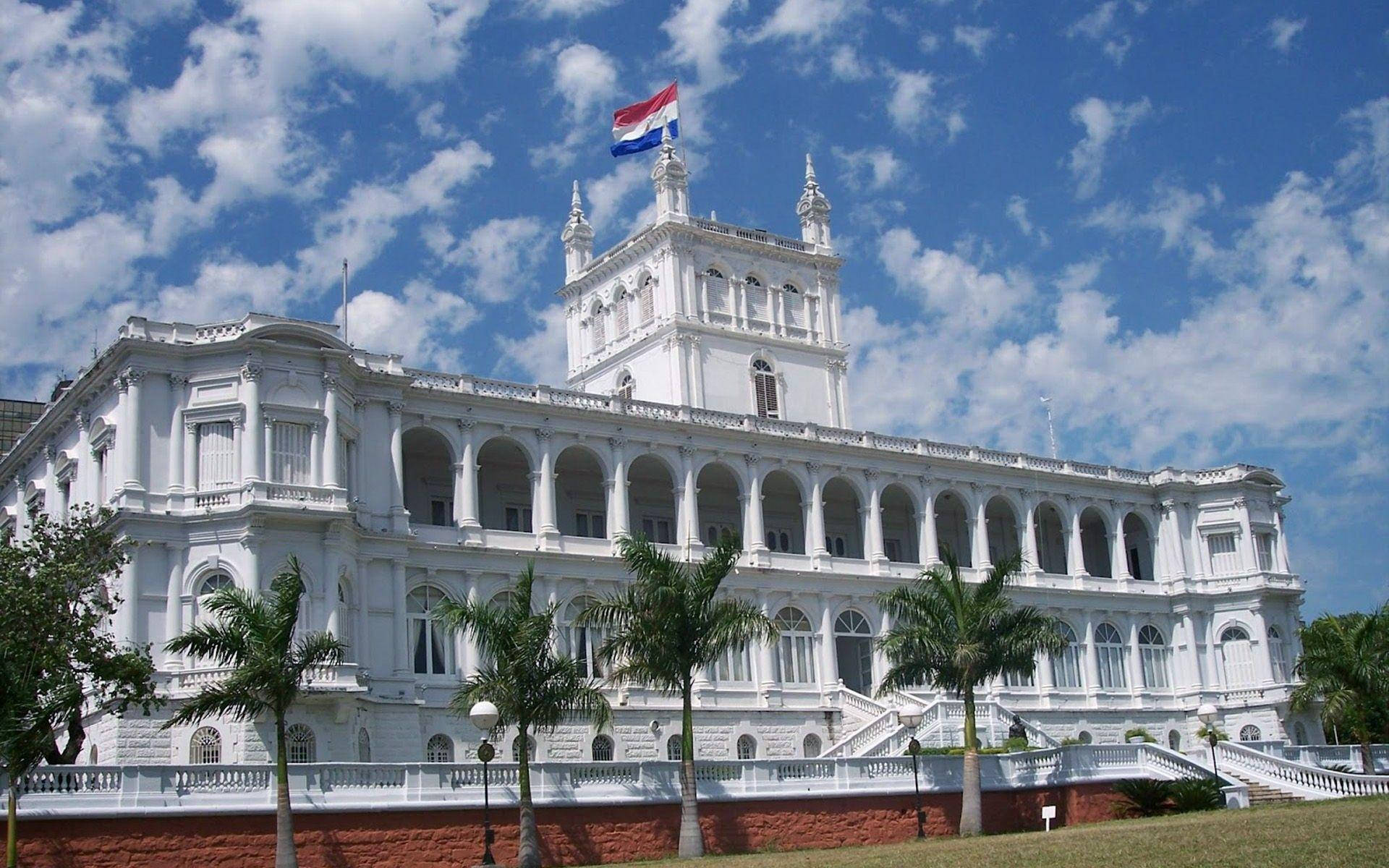 Paraguay 桌布 壁紙 — 一座宏偉的白色殖民風格建築，擁有拱形陽台和頂部的旗幟，周圍環繞著棕櫚樹，背景是明亮的藍天和蓬鬆的白雲。
