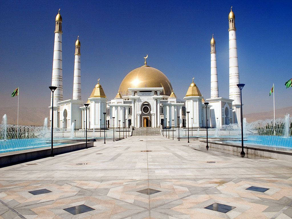 Turkmenistan 桌布 壁紙 — 一座宏偉的白色清真寺，擁有金色圓頂和宣禮塔，矗立在晴朗的藍天下，兩側對稱的噴泉和幾何瓷磚步道。