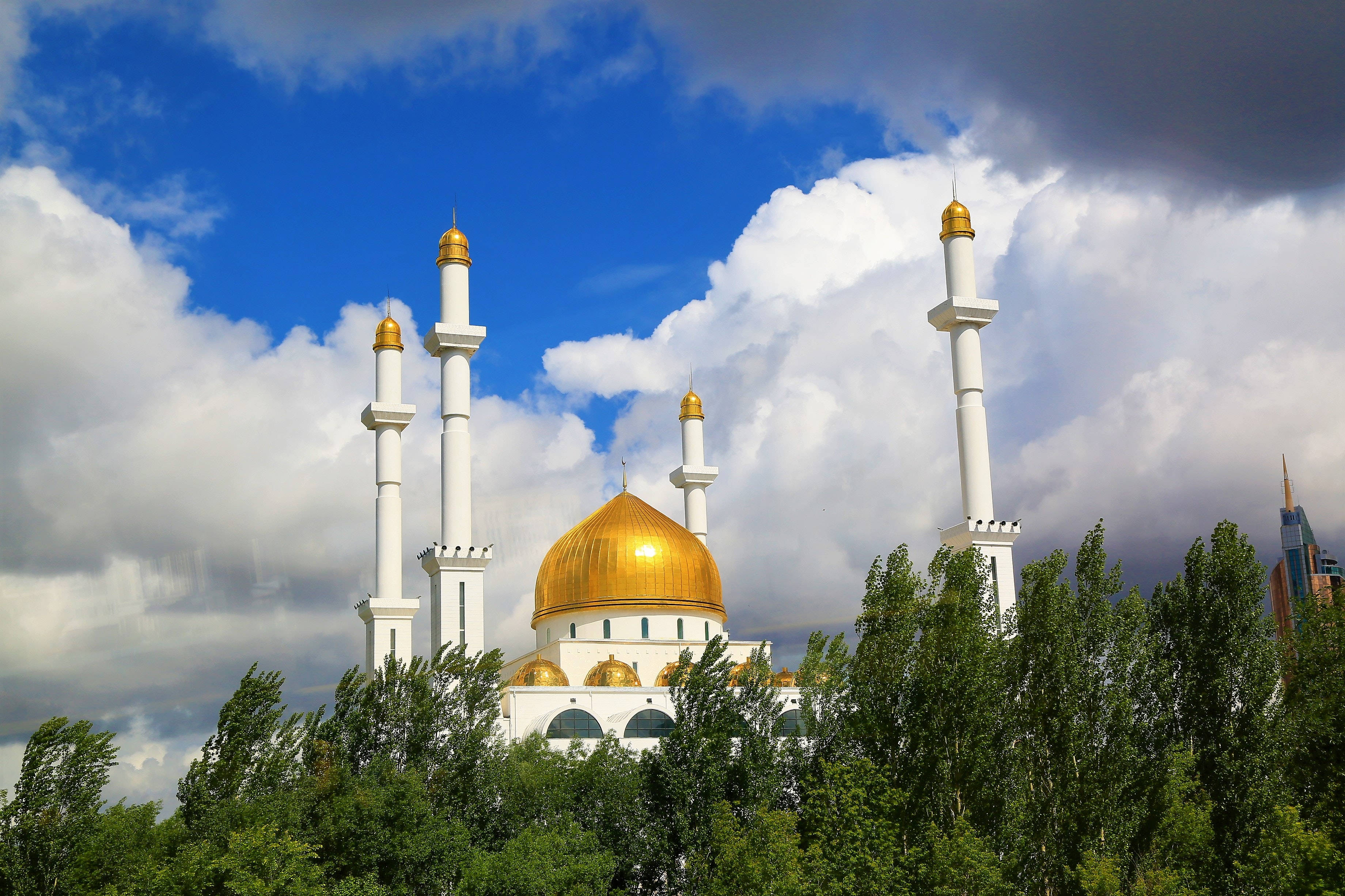Best Islamic 桌布 壁紙 — 一座宏偉的白色清真寺，擁有金色圓頂和宣禮塔，聳立在鬱鬱蔥蔥的綠樹之上，背景是帶有蓬鬆雲朵的戲劇性藍天。