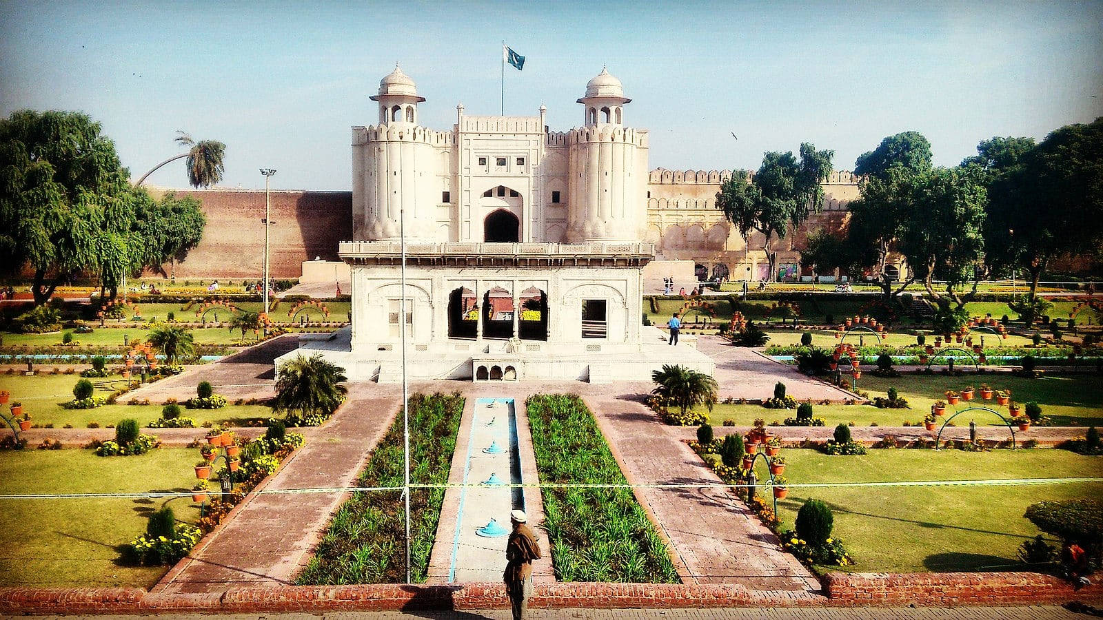 Lahore 桌布 壁紙 — 一座宏偉的白色蒙兀兒風格大門矗立在精心修剪的花園中央，花園有對稱的小徑和充滿活力的綠色草坪，上方是清澈的藍天。