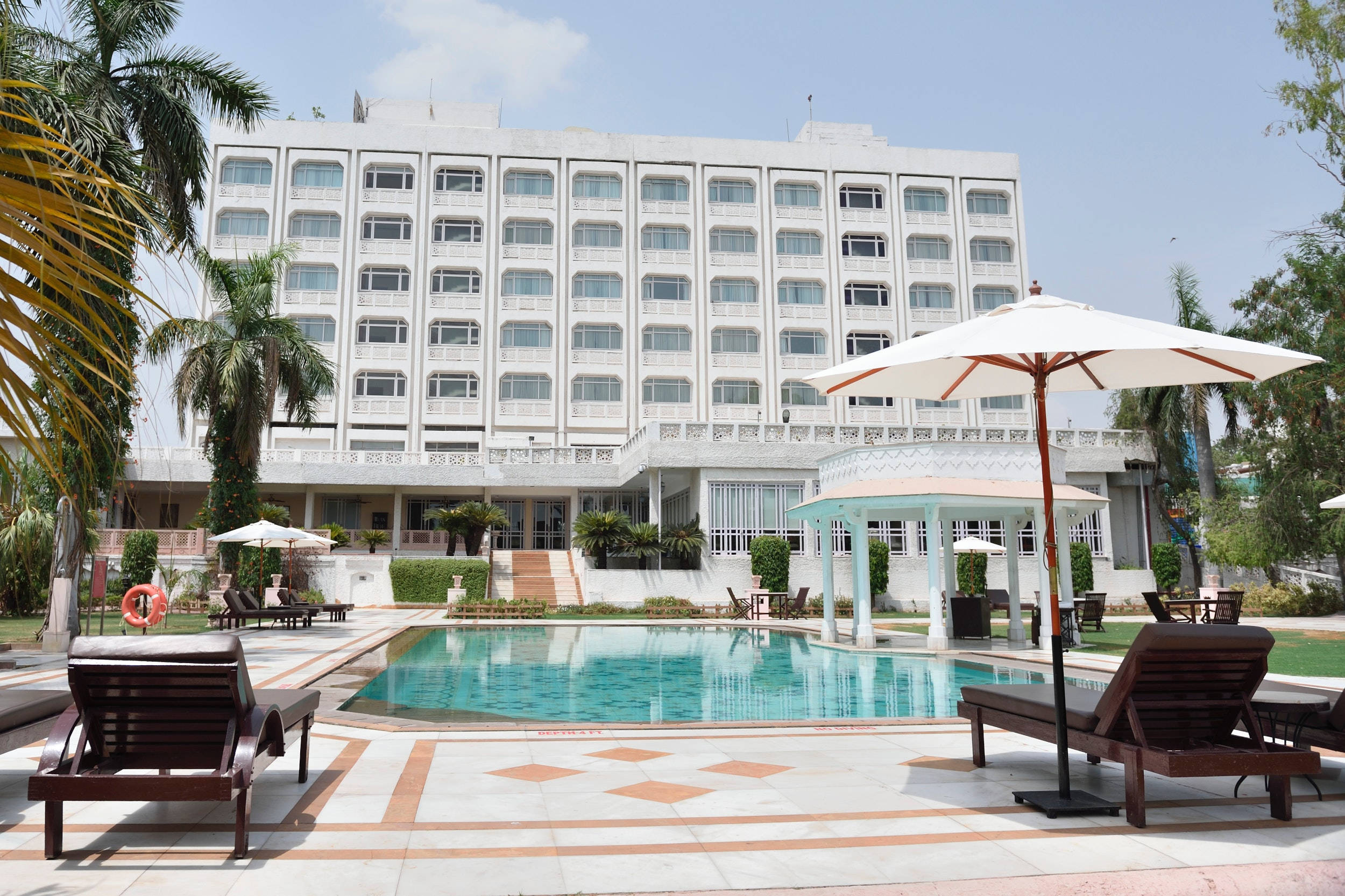 Hotel 桌布 壁紙 — 一座宏偉的白色酒店建築聳立在寧靜的碧藍色泳池上方，周圍環繞著棕櫚樹和躺椅，在明亮的日光下。